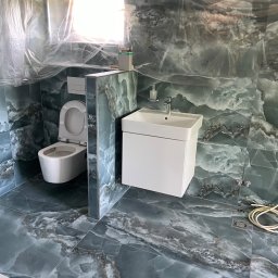 Renewood Piotr Mosór - Wykończona łazienka z toaletą i białą umywalką na tle marmurowych płytek w odcieniach szarości i zieleni. Okno zakryte folią ochronną. Widoczne przewody.