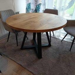 Renewood Piotr Mosór - Okrągły stół z blatem z jasnego drewna i czarną, krzyżową podstawą, otoczony szarymi krzesłami na tle wzorzystej zasłony i szarego dywanu.