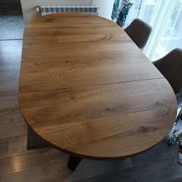 Renewood Piotr Mosór - Rozkładany, owalny stół z blatem z jasnego drewna dębowego, z widocznymi słojami, na ciemnej nodze. W tle krzesła i okno z zasłonami.