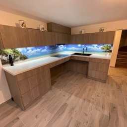 Renewood Piotr Mosór - Nowoczesna kuchnia na wymiar z drewnianymi frontami i fototapetą z motywem plaży. Uwagę przykuwają detale: kubki na szafkach i drewniane schody w tle.