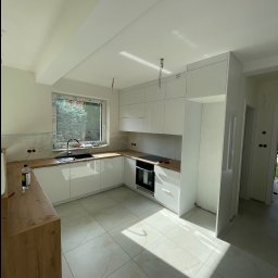 Renewood Piotr Mosór - Jasna kuchnia z białymi szafkami i drewnianym blatem, widok na okno i fragment podłogi z płytek. W tle widoczne niedokończone prace wykończeniowe.