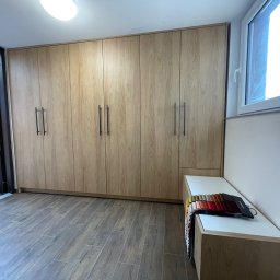 Renewood Piotr Mosór - Jasna szafa na wymiar z drewnianymi frontami i czarnymi uchwytami, dopasowana do wnętrza. Na blacie próbki materiałów. Widok z perspektywy podłogi.