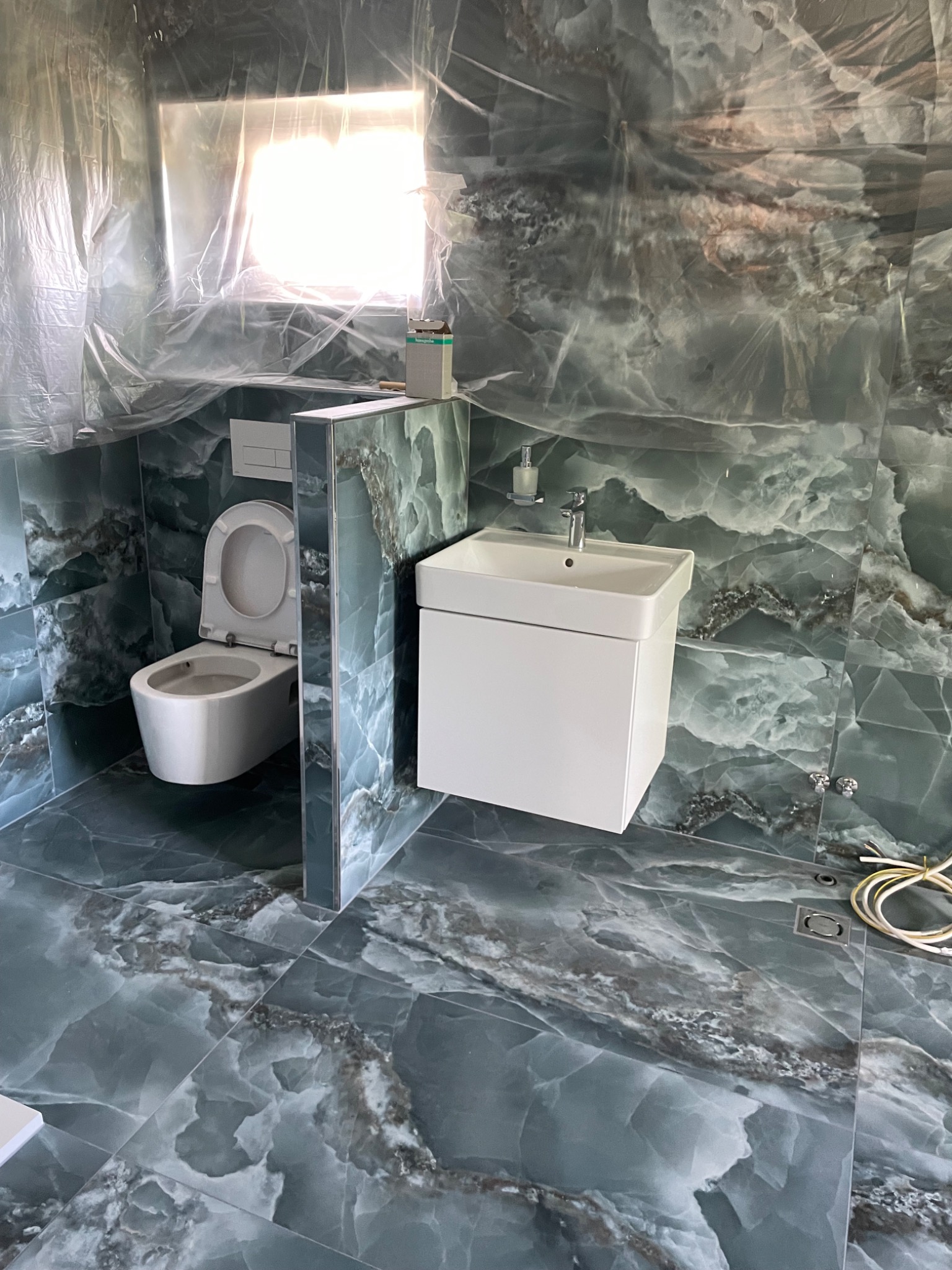 Wykończona łazienka z toaletą i białą umywalką na tle marmurowych płytek w odcieniach szarości i zieleni. Okno zakryte folią ochronną. Widoczne przewody.
