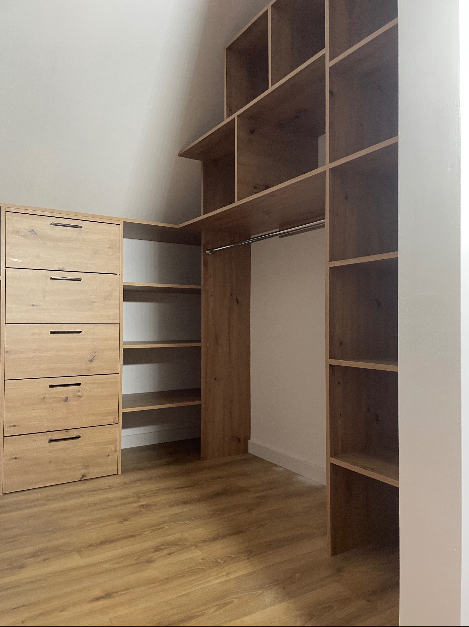 Jasna, drewniana szafa wnękowa z szufladami i półkami, dopasowana do skosu dachu na poddaszu. Widoczny drążek na ubrania i drewniana podłoga.