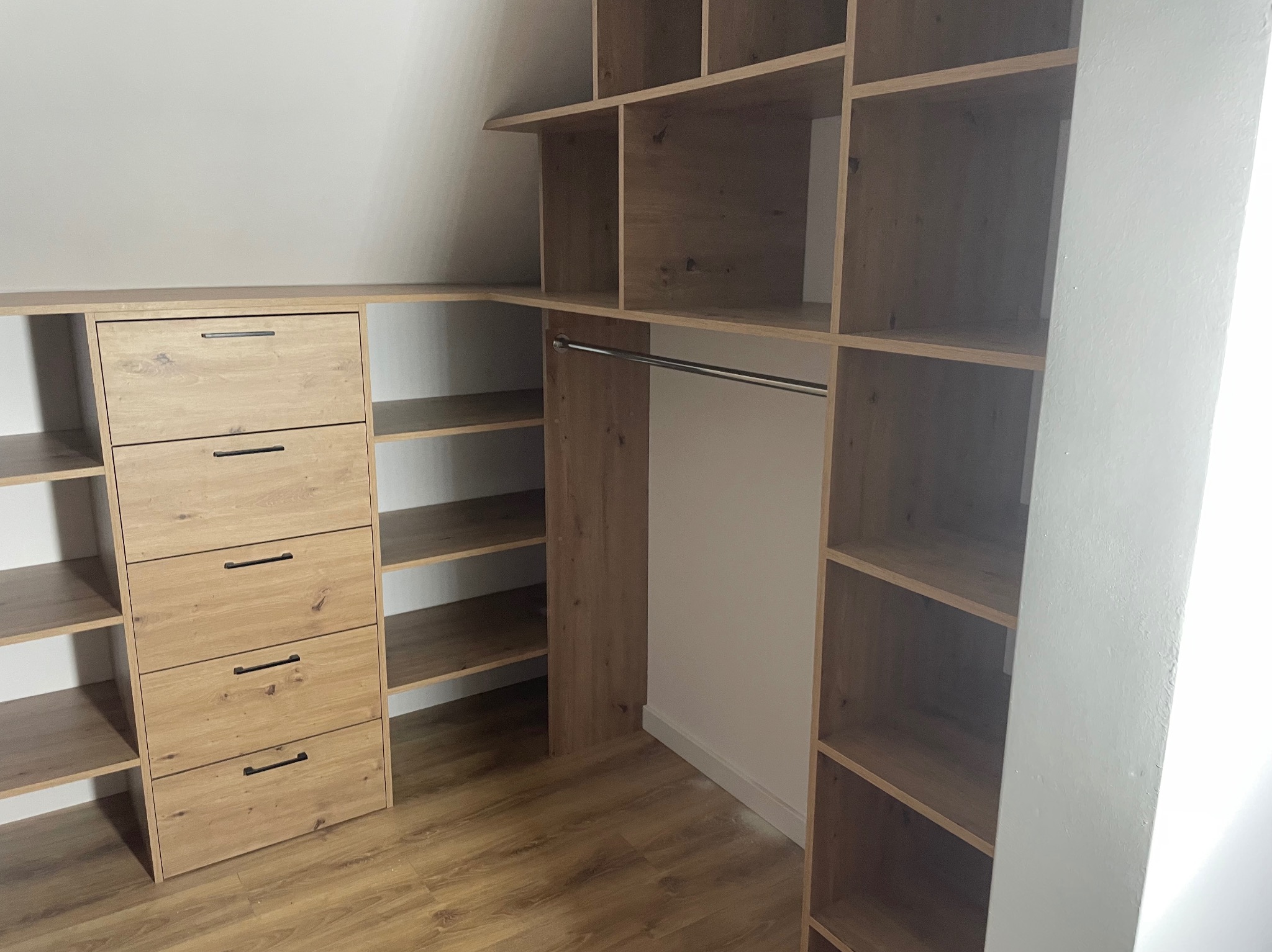 Drewniana szafa wnękowa z szufladami i półkami, wykończona w jasnym odcieniu drewna. Widoczny drążek na ubrania. Ustawiona na poddaszu ze skosem.