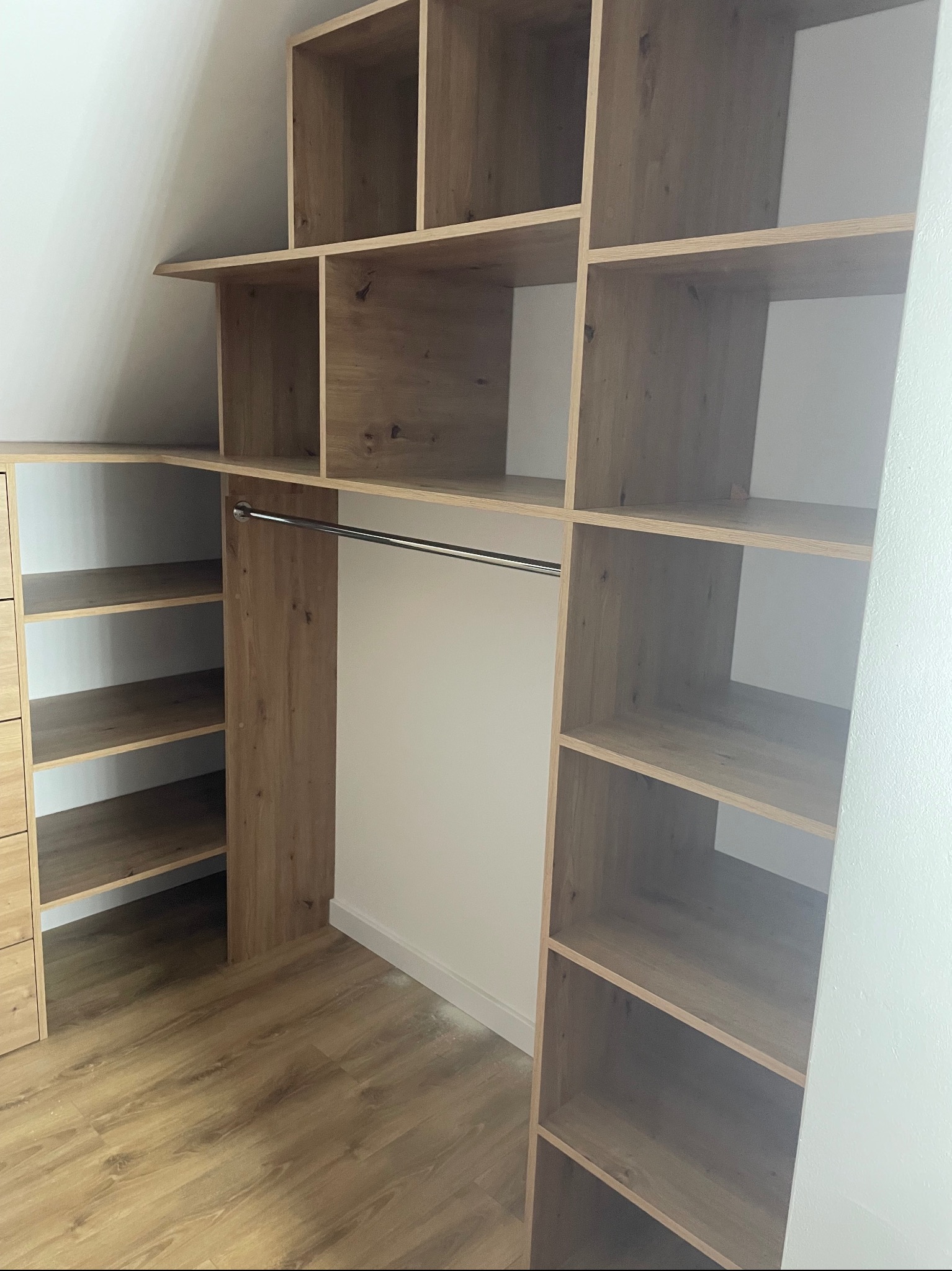 Drewniana szafa wnękowa z półkami i drążkiem na ubrania, dopasowana do skosu dachu. Widoczne drewniane wykończenie i jasne wnętrze szafy.