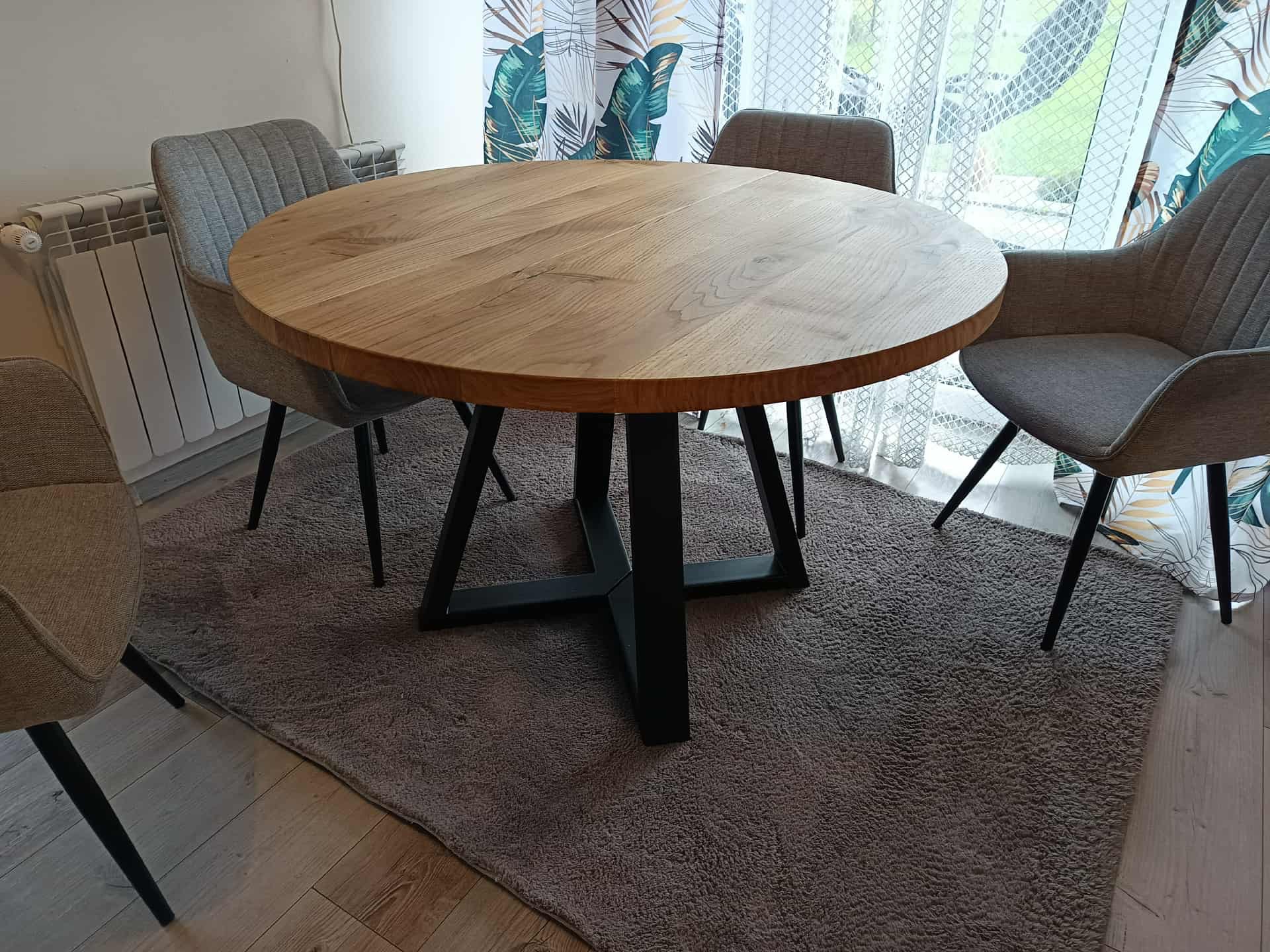 Okrągły stół z blatem z jasnego drewna i czarną, krzyżową podstawą, otoczony szarymi krzesłami na tle wzorzystej zasłony i szarego dywanu.