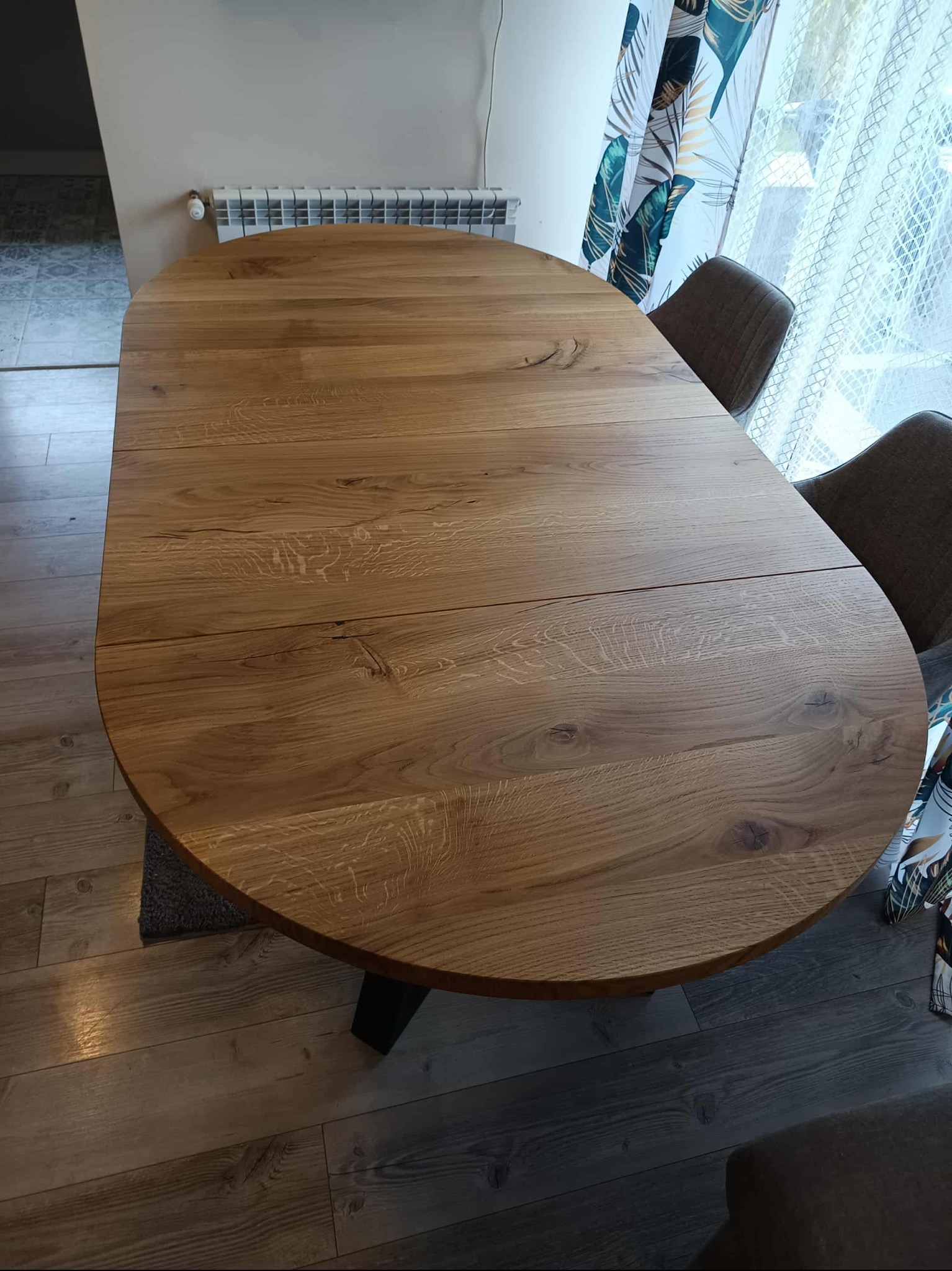 Rozkładany, owalny stół z blatem z jasnego drewna dębowego, z widocznymi słojami, na ciemnej nodze. W tle krzesła i okno z zasłonami.