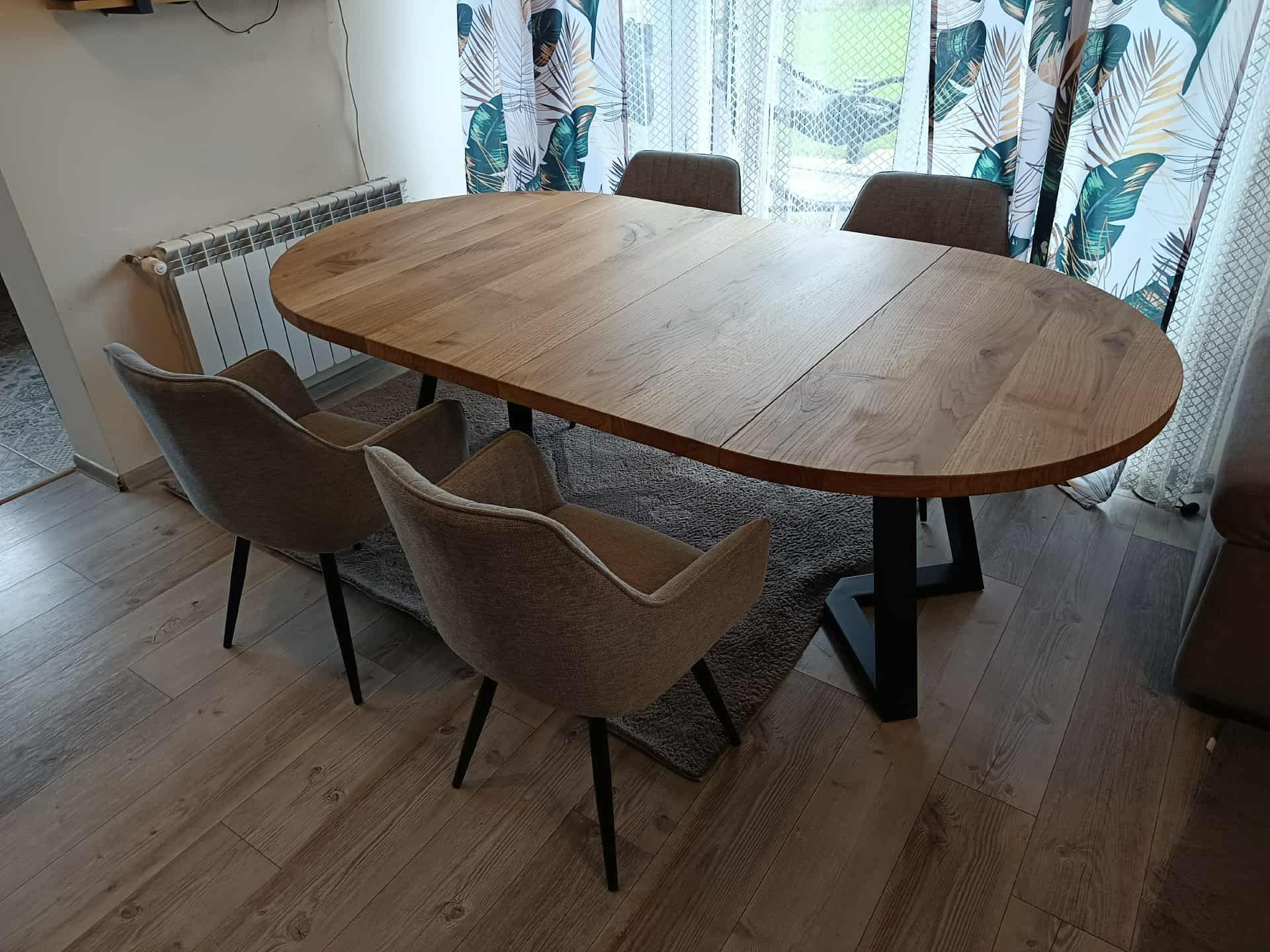 Rozkładany stół z blatem z jasnego drewna i czarnymi nogami, otoczony czterema krzesłami w odcieniach szarości, na tle okna z wzorzystymi zasłonami.