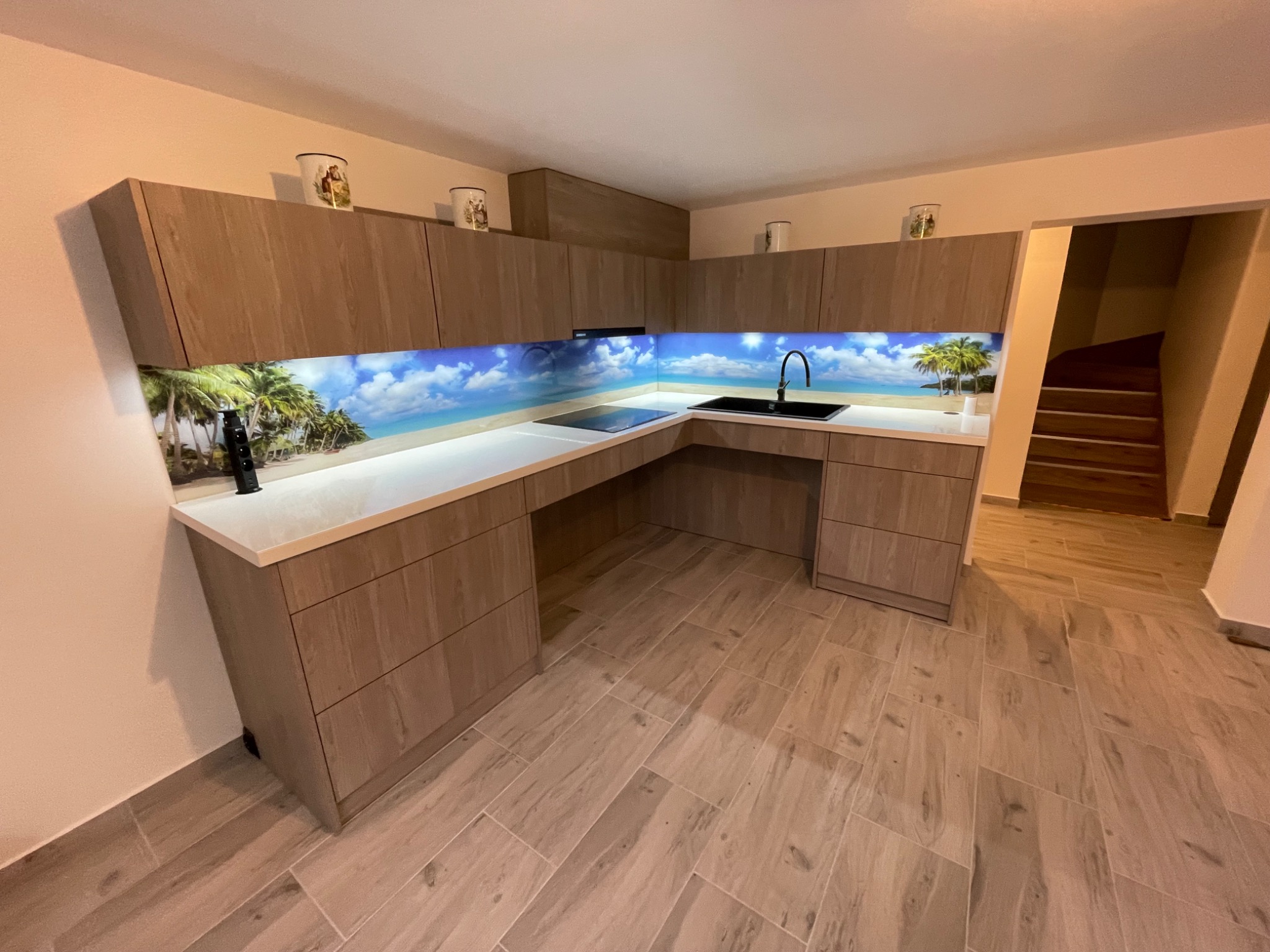 Nowoczesna kuchnia na wymiar z drewnianymi frontami i fototapetą z motywem plaży. Uwagę przykuwają detale: kubki na szafkach i drewniane schody w tle.