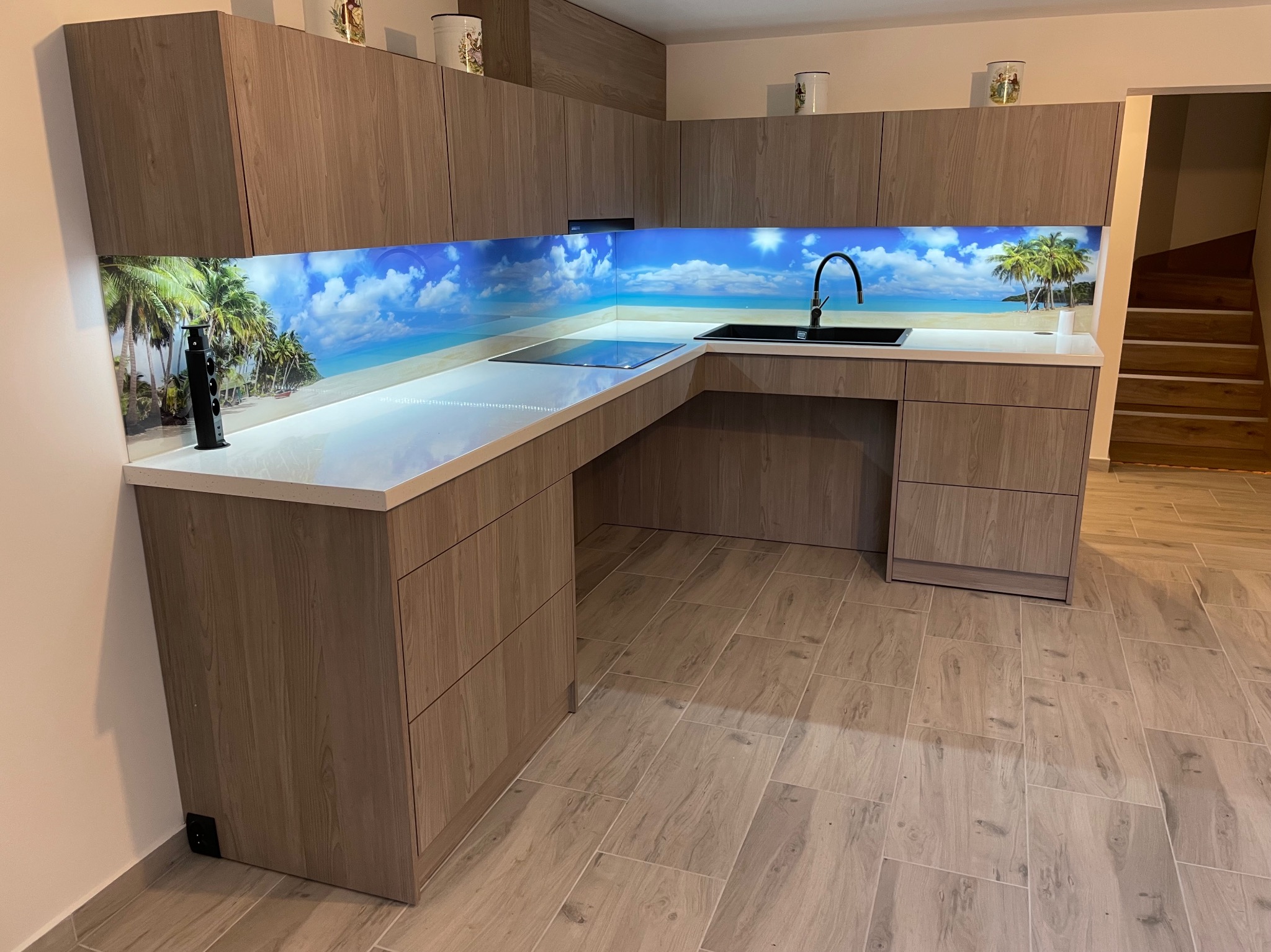 Nowoczesna kuchnia w odcieniach drewna z blatem i panelem nad blatem z motywem plaży. Widoczne szafki górne i dolne, zlew oraz fragment schodów w tle.