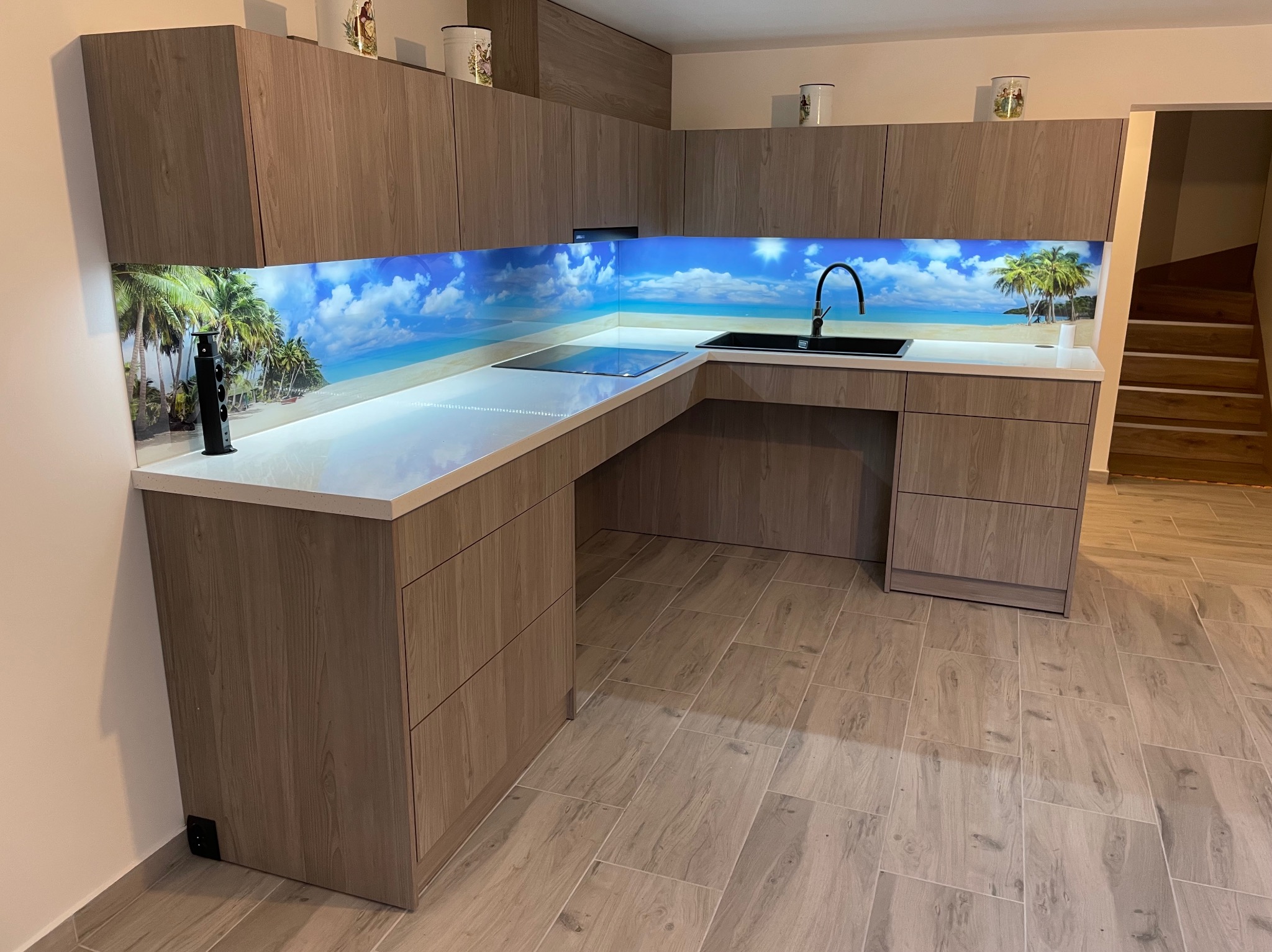 Nowoczesna kuchnia w kształcie litery L z drewnianymi frontami i blatem w kolorze bieli. Nad blatem panel z motywem plaży. Widoczne schody w tle.