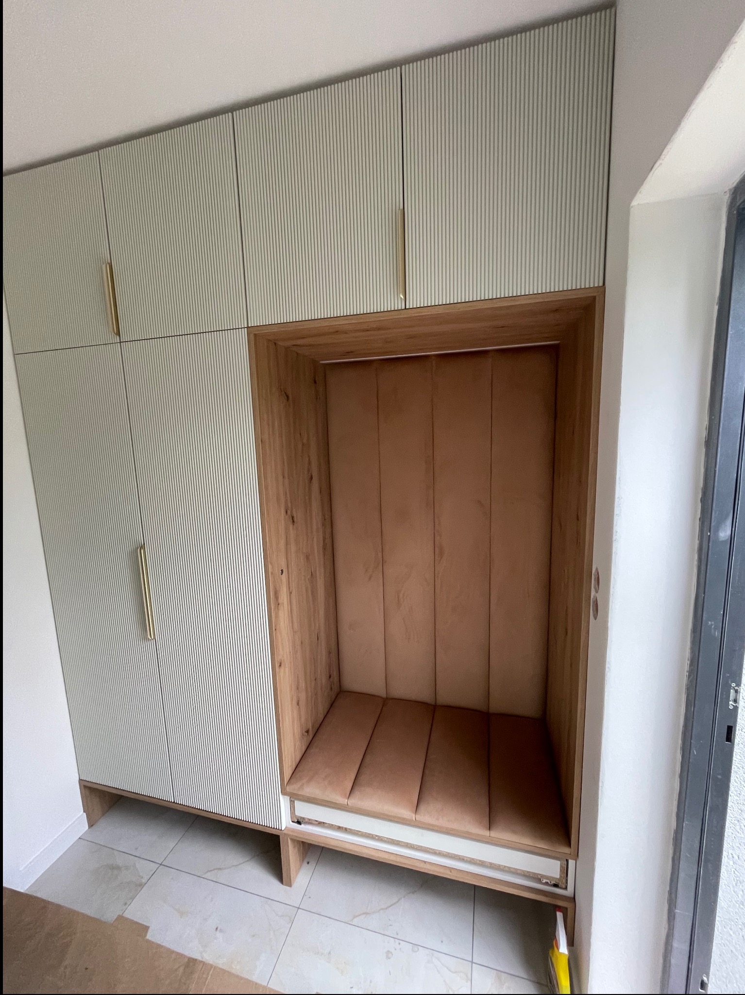 Biało-zielona szafa z ryflowanymi frontami i złotymi uchwytami, z wnęką wykończoną drewnem i tapicerowanym siedziskiem w kolorze beżowym.