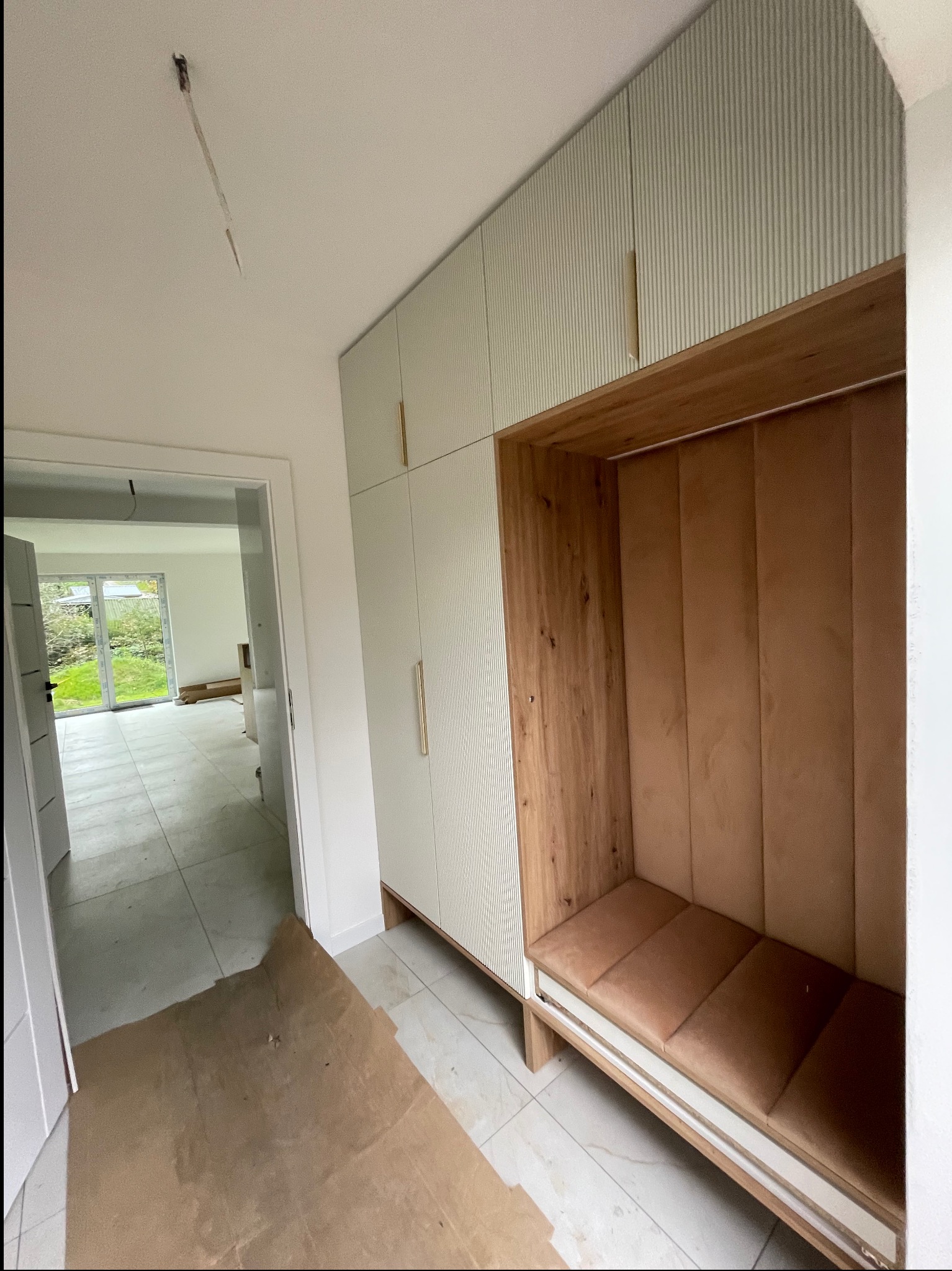 Nowoczesna szafa wnękowa z siedziskiem w przedpokoju. Jasne fronty szafek z ryflowanym wzorem, drewniane wykończenie siedziska i oparcia. Widok na wnętrze domu.