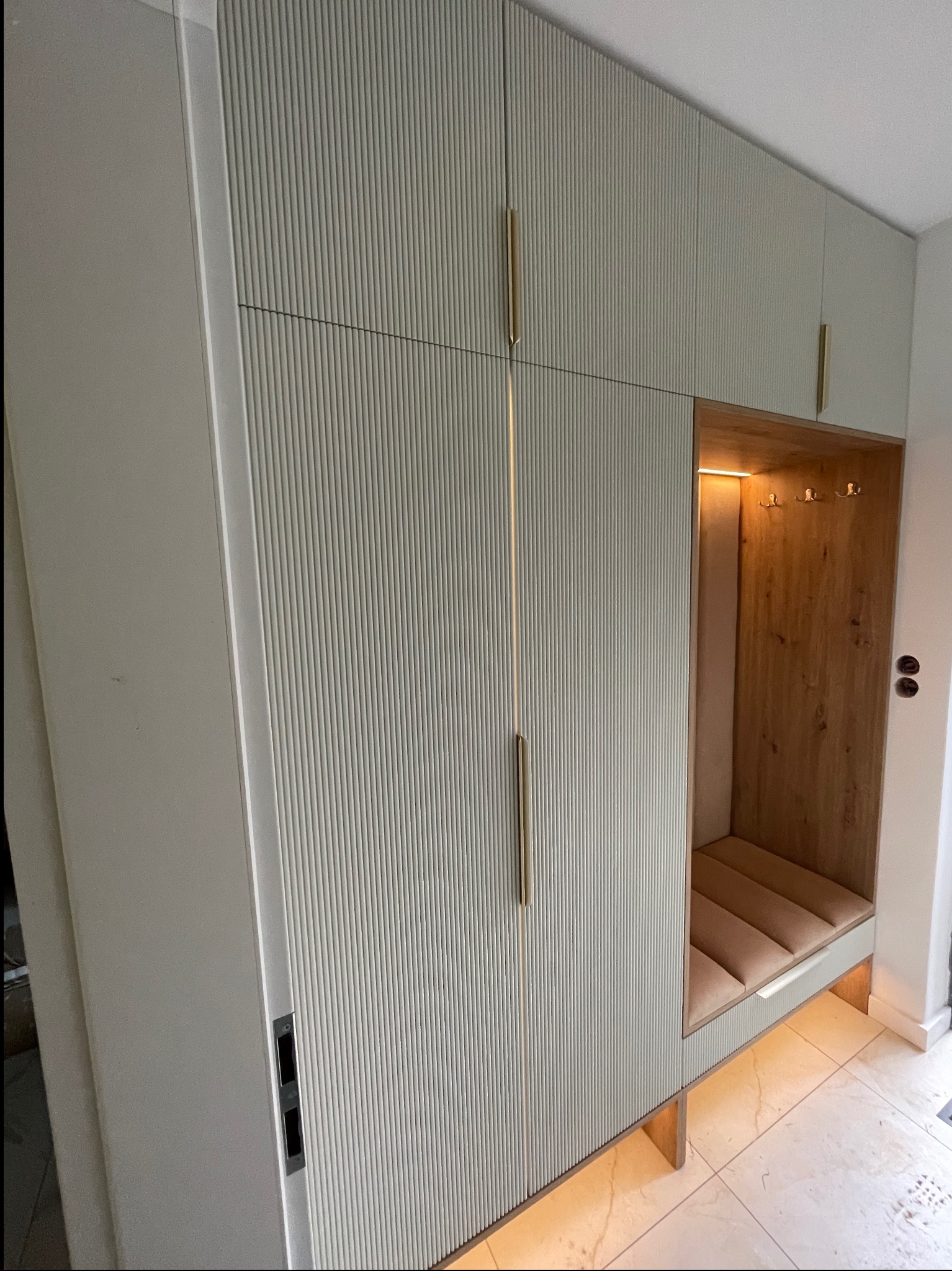 Jasna szafa wnękowa z ryflowanymi frontami i złotymi uchwytami, z wbudowanym siedziskiem z oświetleniem LED. Drewniane wykończenie wnęki z haczykami.