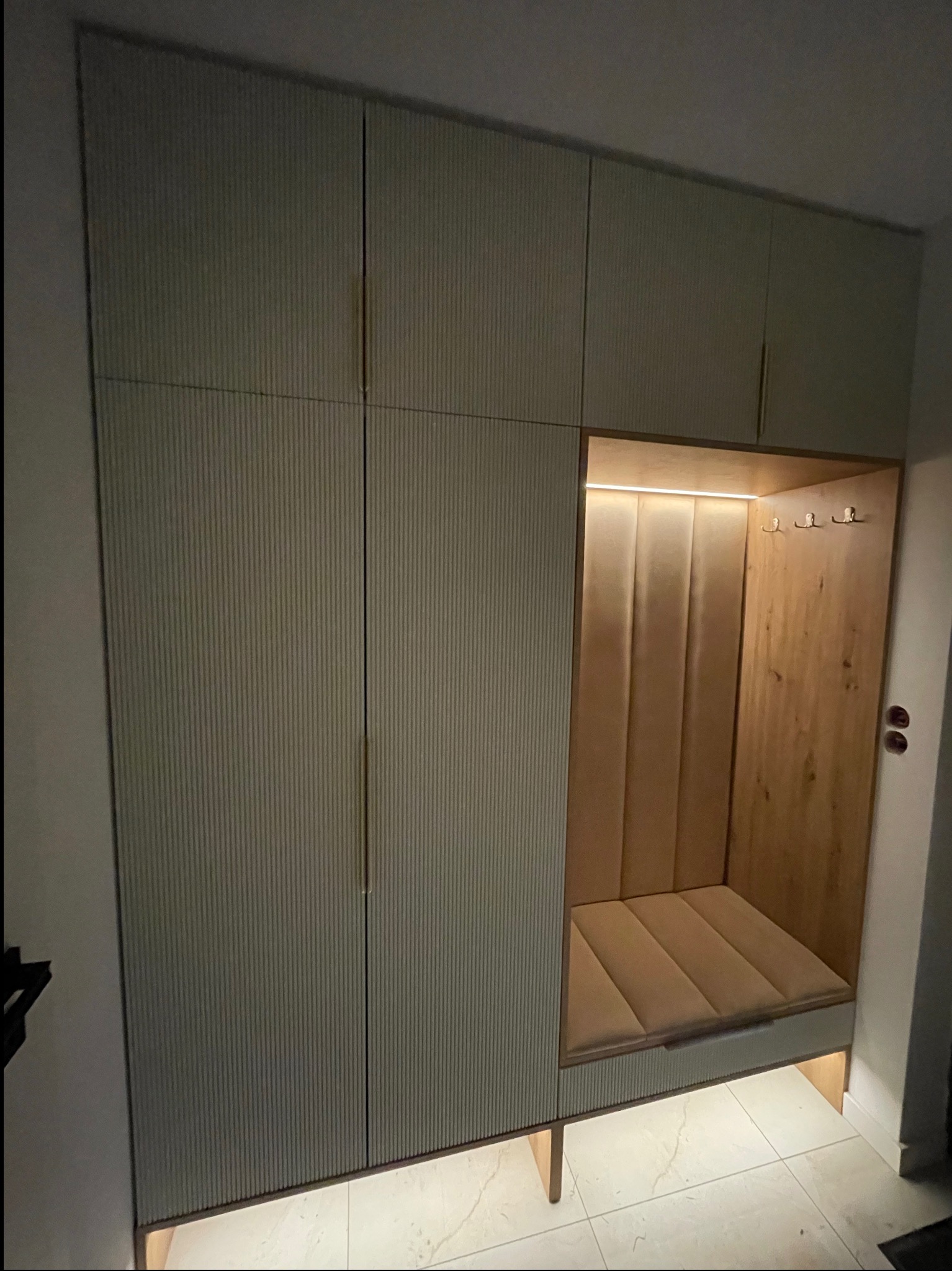Nowoczesna szafa wnękowa z siedziskiem i wieszakami, oświetlona LED, z ryflowanymi frontami w odcieniach beżu i drewna. Minimalistyczny design, jasna podłoga.