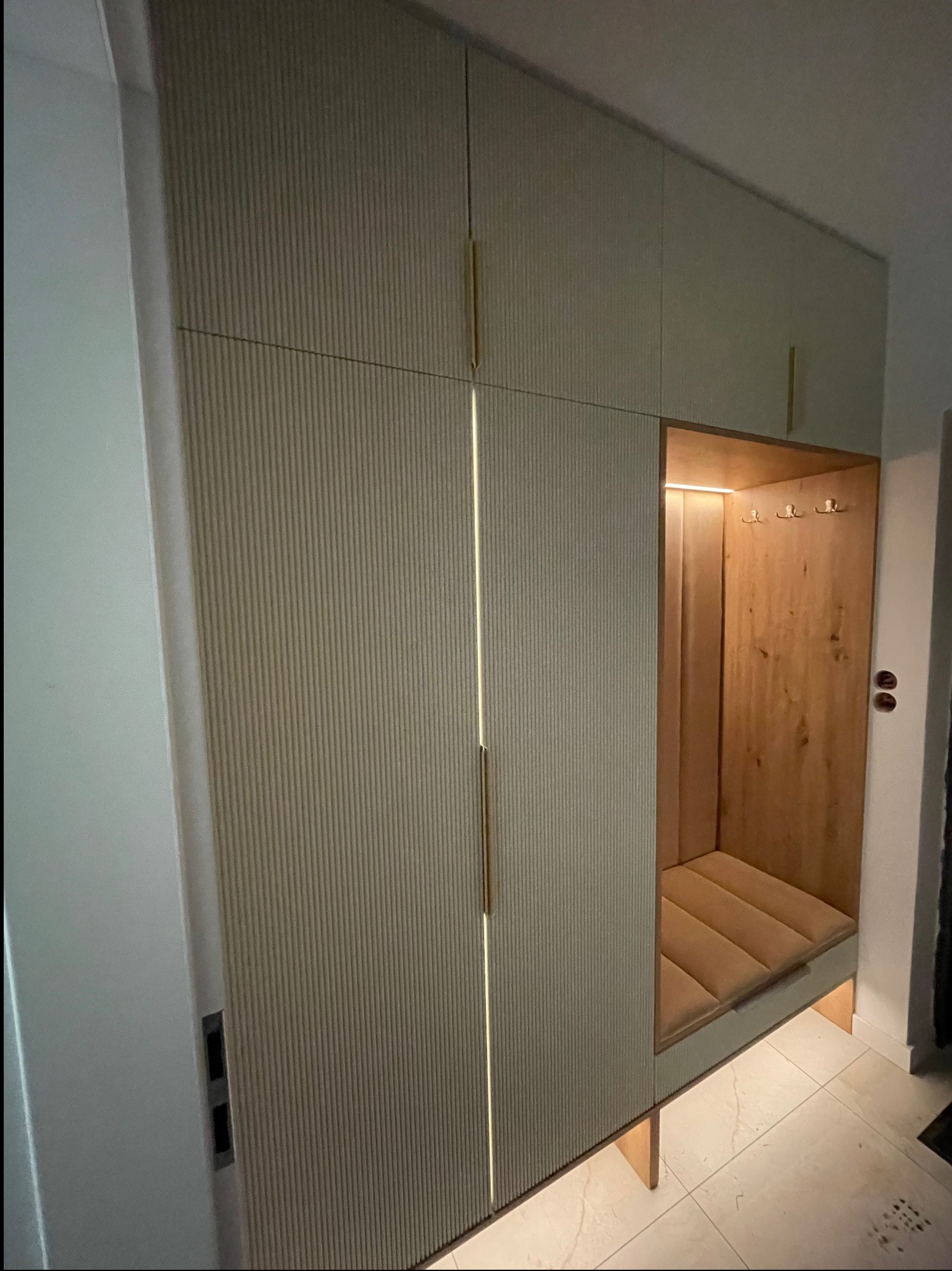 Nowoczesna szafa wnękowa z siedziskiem i oświetleniem LED. Fronty ryflowane w odcieniach beżu, złote uchwyty. Wnętrze siedziska wykończone drewnem i tapicerowane.