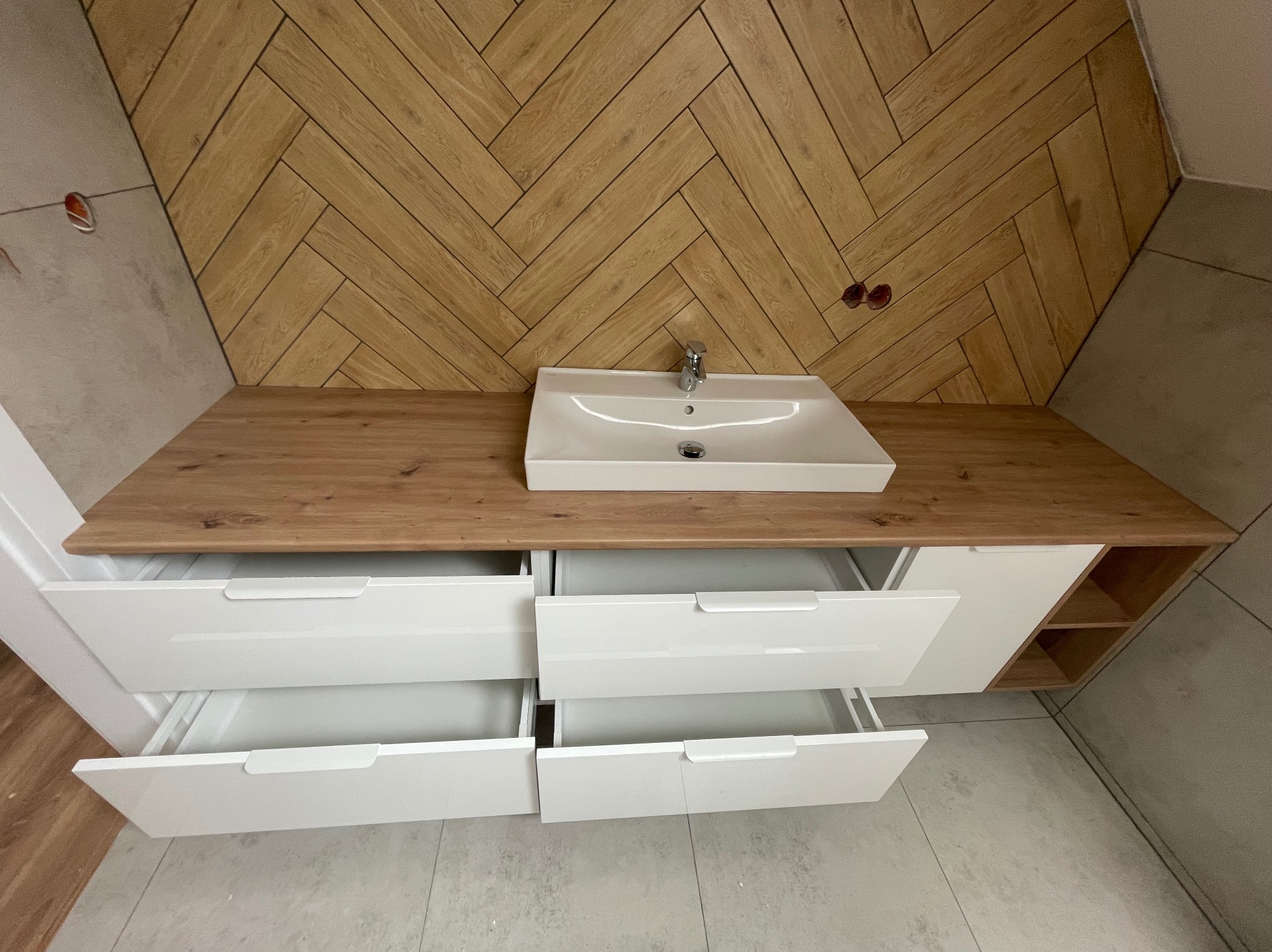 Nowoczesna łazienka z drewnianą ścianą w jodełkę, blatem z umywalką i białymi szafkami z otwartymi szufladami. Widok z góry podkreśla unikalny wzór drewna.