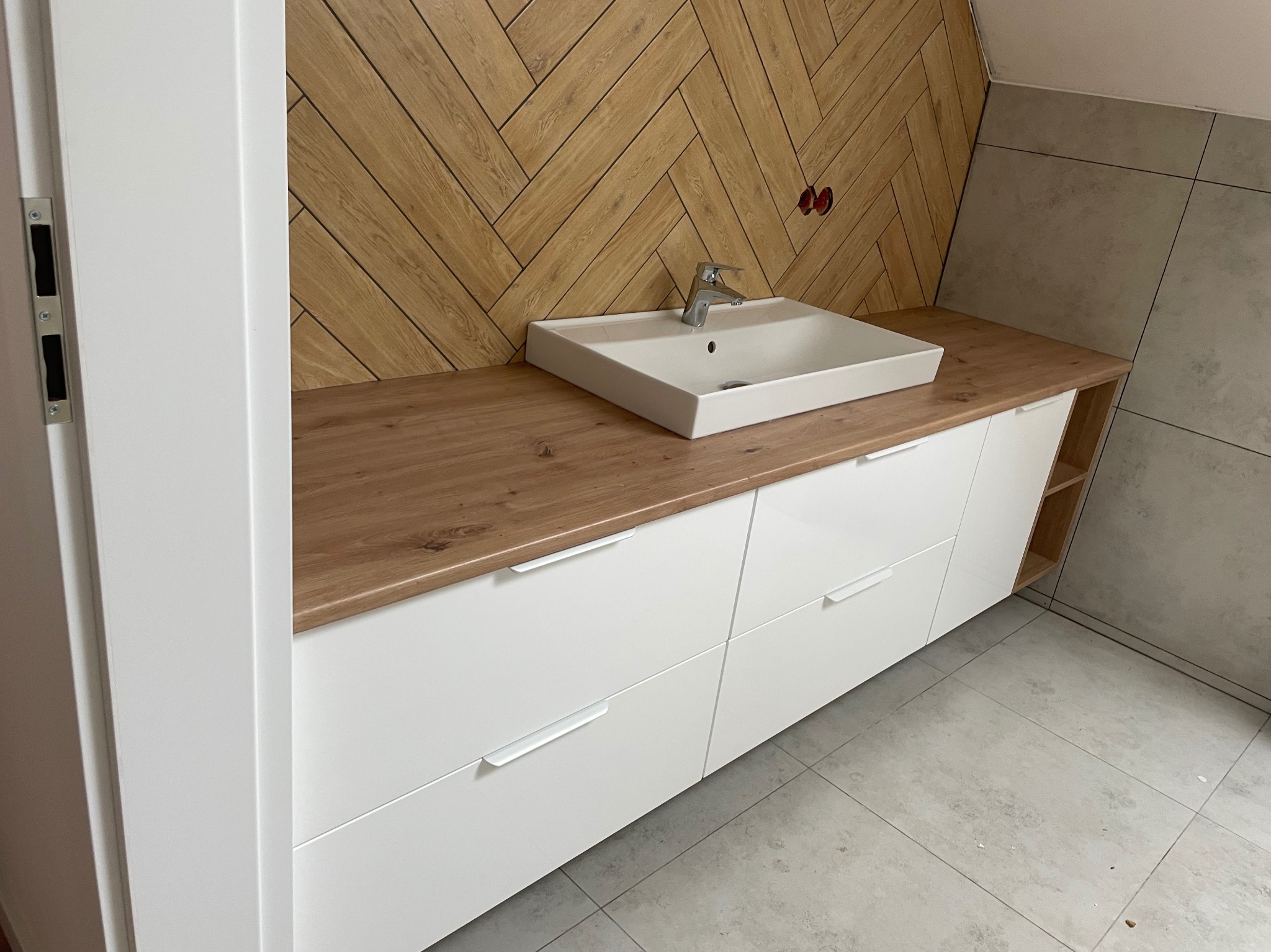 Nowoczesna szafka łazienkowa z białymi frontami i drewnianym blatem, umywalka nablatowa. Ściana wykończona drewnem ułożonym w jodełkę i płytkami. Minimalistyczny design.