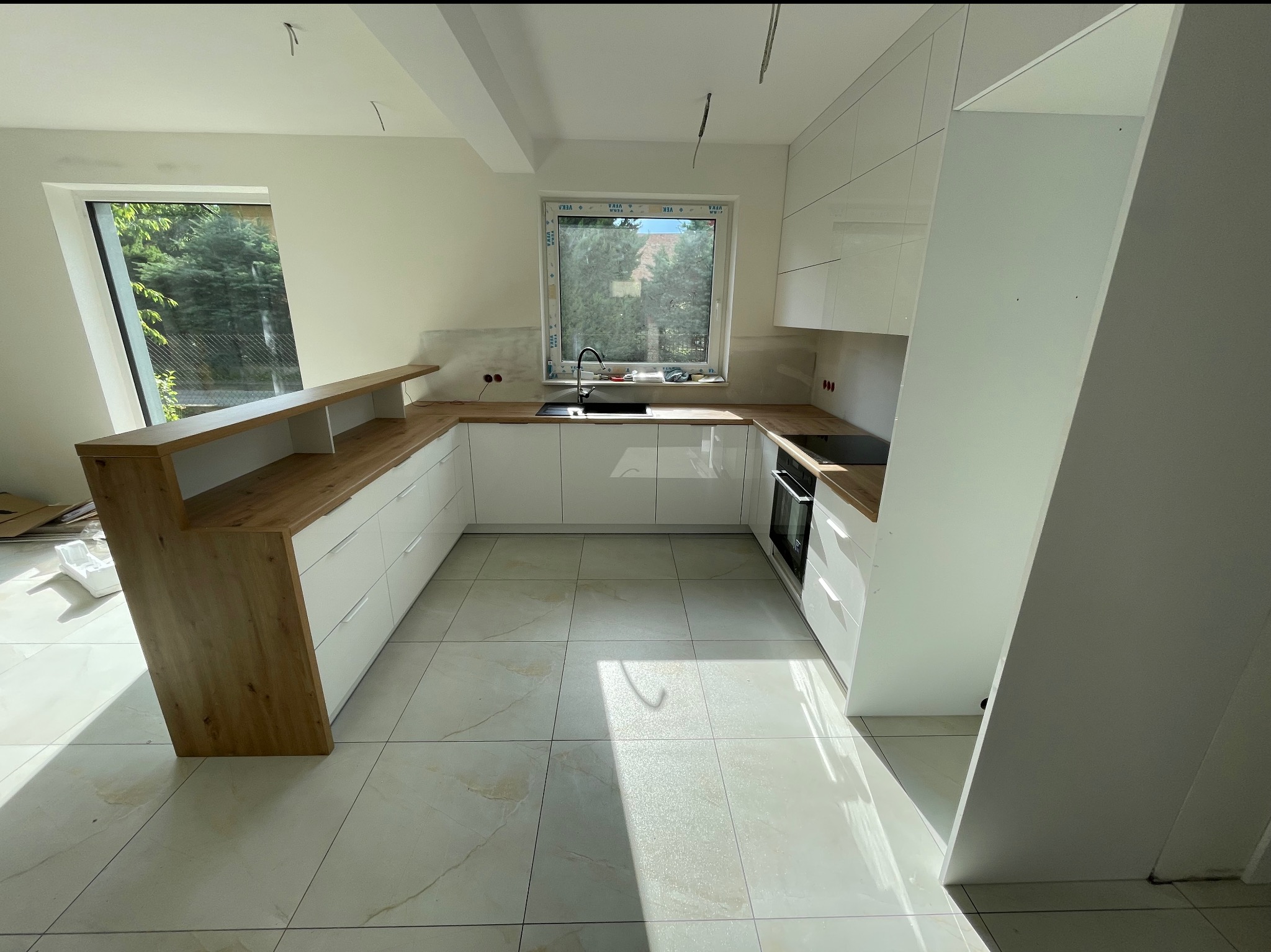 Nowoczesna kuchnia w kształcie litery U z białymi szafkami i drewnianym blatem, oświetlona naturalnym światłem z okna. Widoczne płytki podłogowe w jasnym odcieniu.