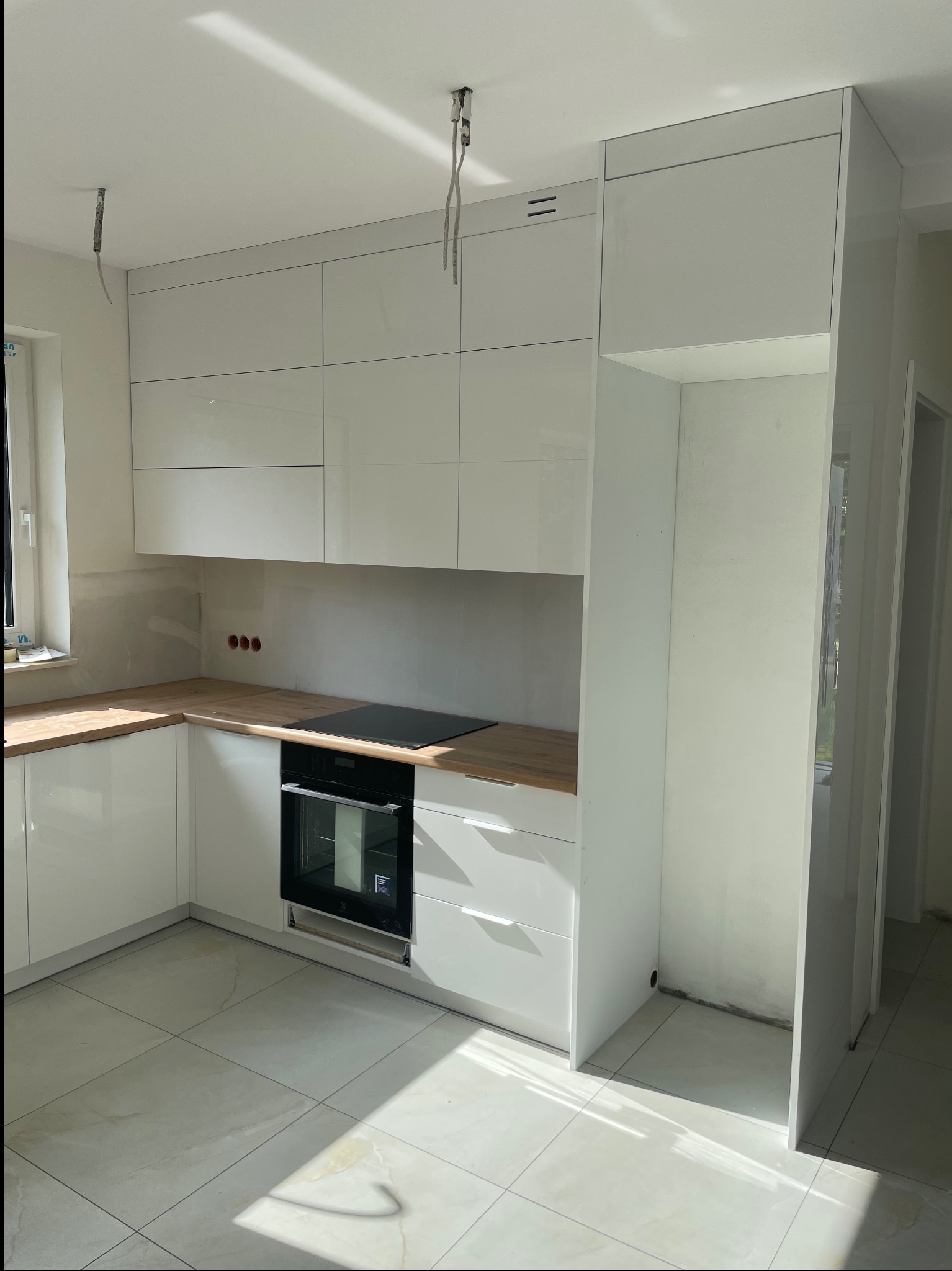 Jasna kuchnia z białymi, lakierowanymi frontami i drewnianym blatem. Widoczne niedokończone prace wykończeniowe, takie jak wystające kable i brak listew przypodłogowych.