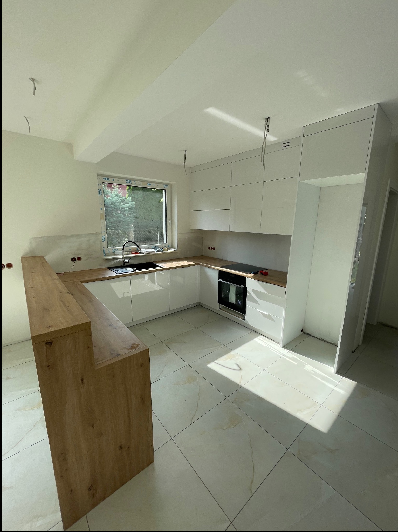Jasna kuchnia z drewnianym blatem i białymi szafkami, w trakcie montażu. Widoczne niedokończone ściany i wystające kable. Nowoczesny design i marmurowe płytki podłogowe.