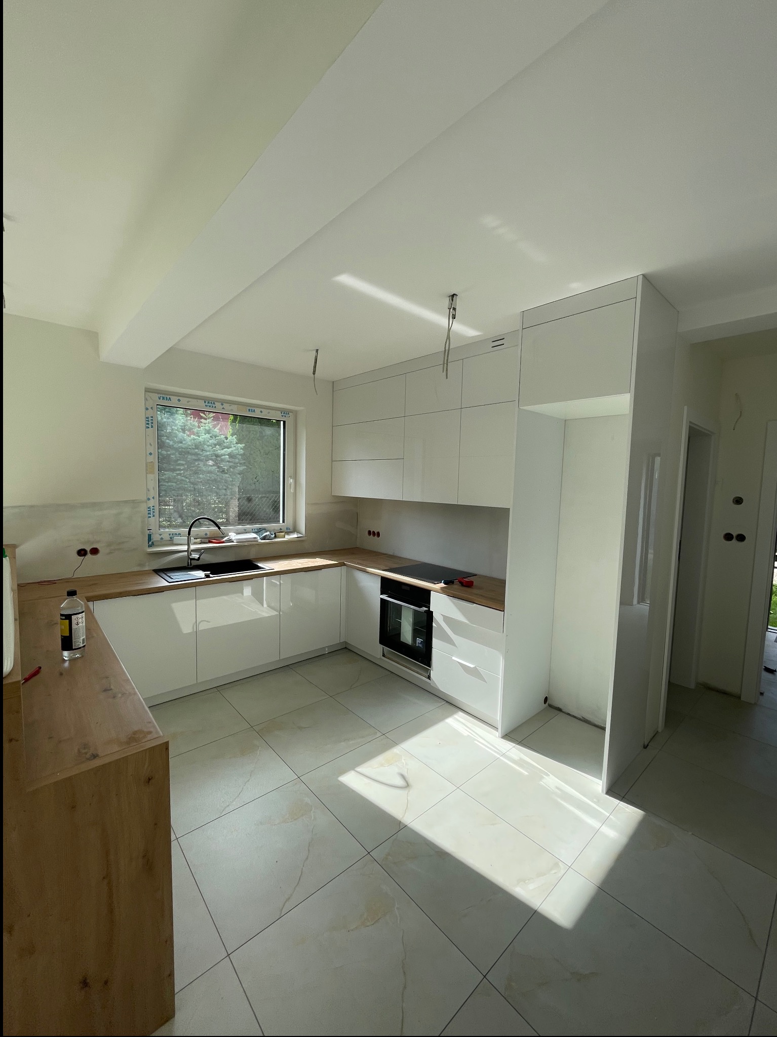 Jasna kuchnia z białymi szafkami i drewnianym blatem, widok na okno i fragment podłogi z płytek. W tle widoczne niedokończone prace wykończeniowe.