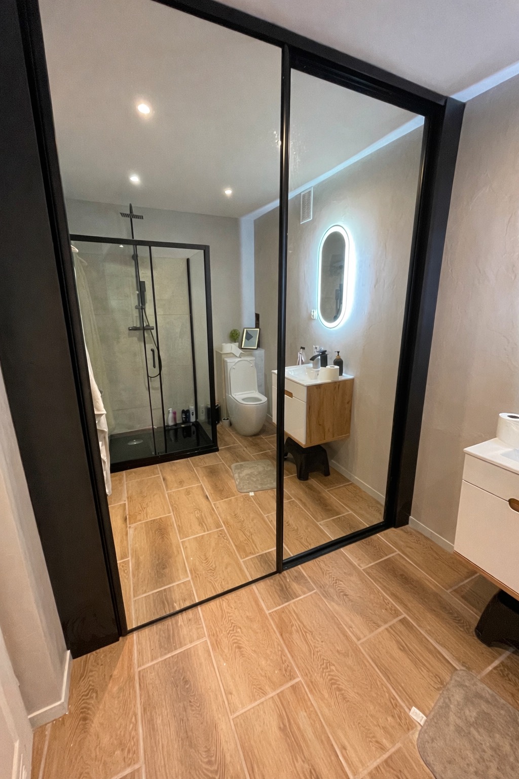 Nowoczesna łazienka za przesuwnymi drzwiami z czarną ramą. Widoczny prysznic typu walk-in, toaleta, umywalka z podświetlanym lustrem i drewnopodobne płytki na podłodze.