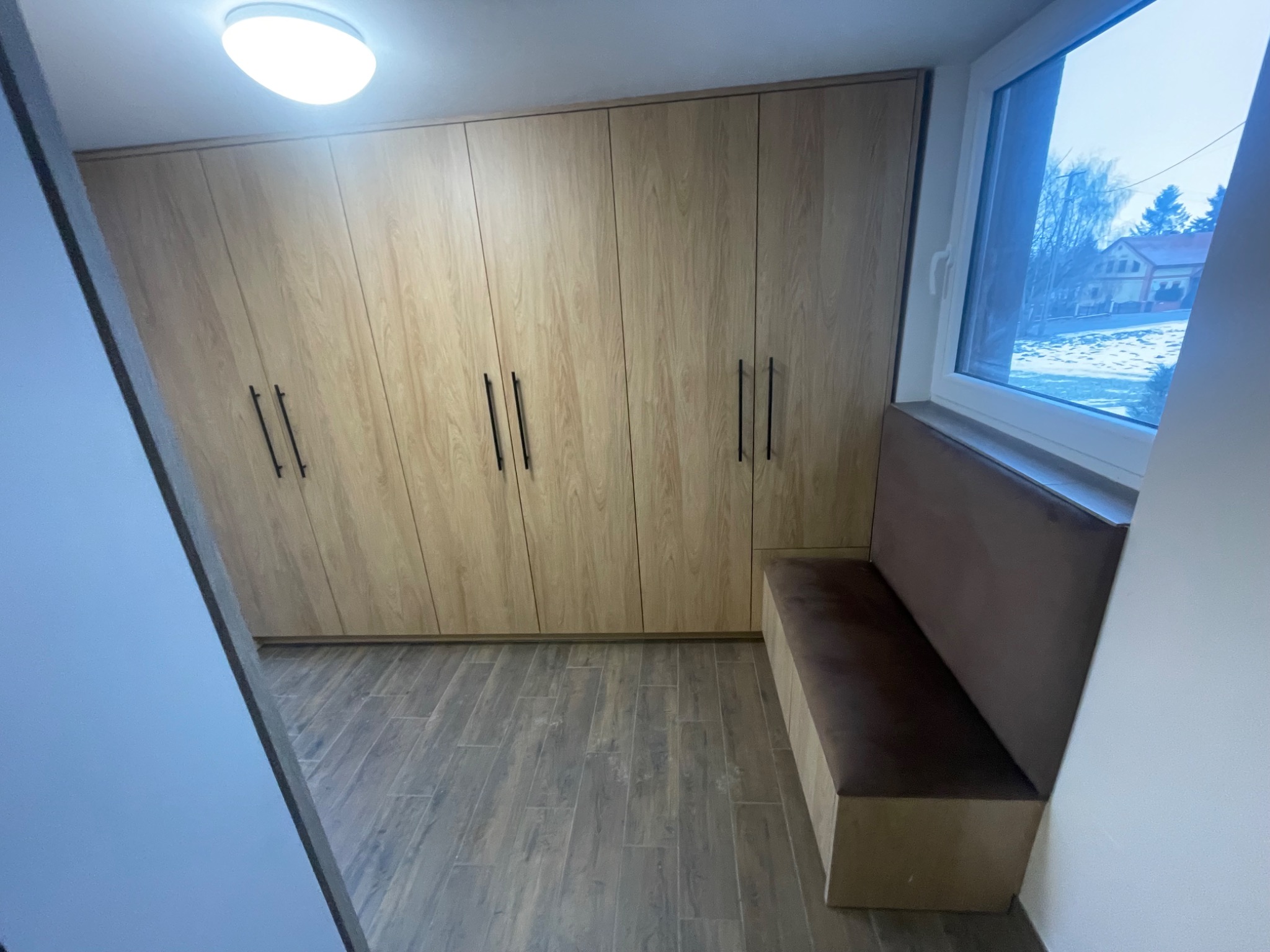 Drewniana szafa na wymiar z czarnymi uchwytami, dopasowana do wnęki z siedziskiem przy oknie. Podłoga wyłożona płytkami imitującymi drewno. Widok na zimowy krajobraz.