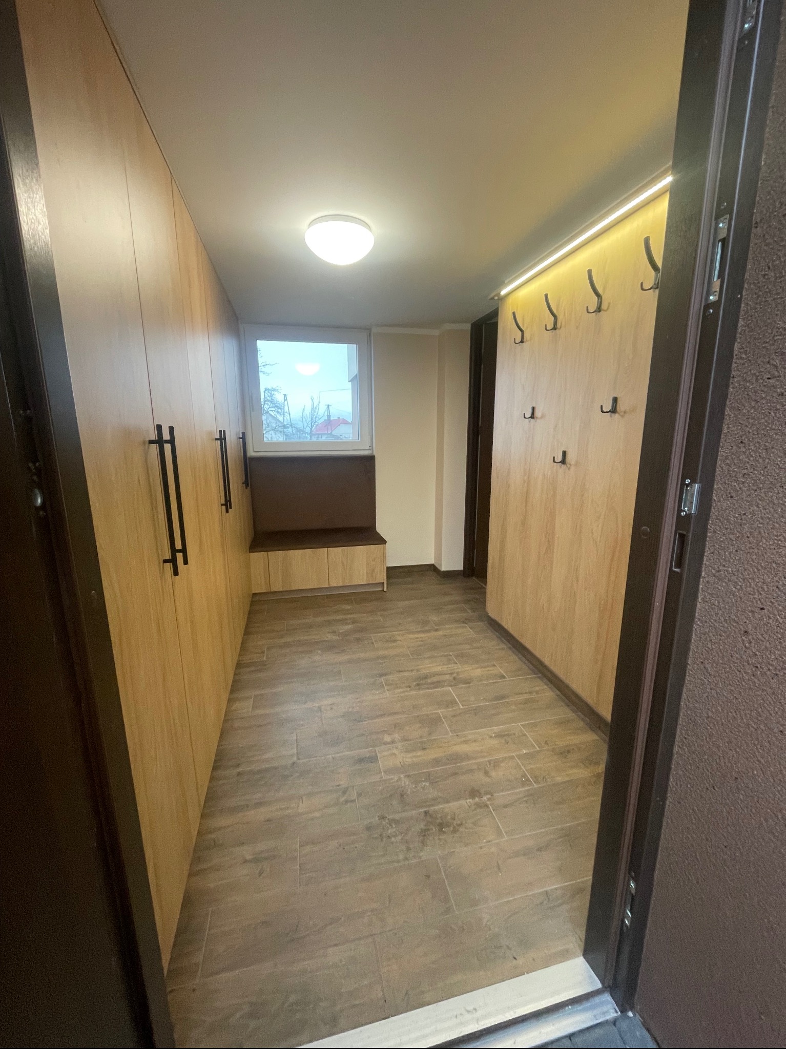 Funkcjonalna zabudowa przedpokoju z drewnianymi szafami na wymiar, wnęką z siedziskiem i wieszakami. Oświetlenie LED podkreśla nowoczesny design.