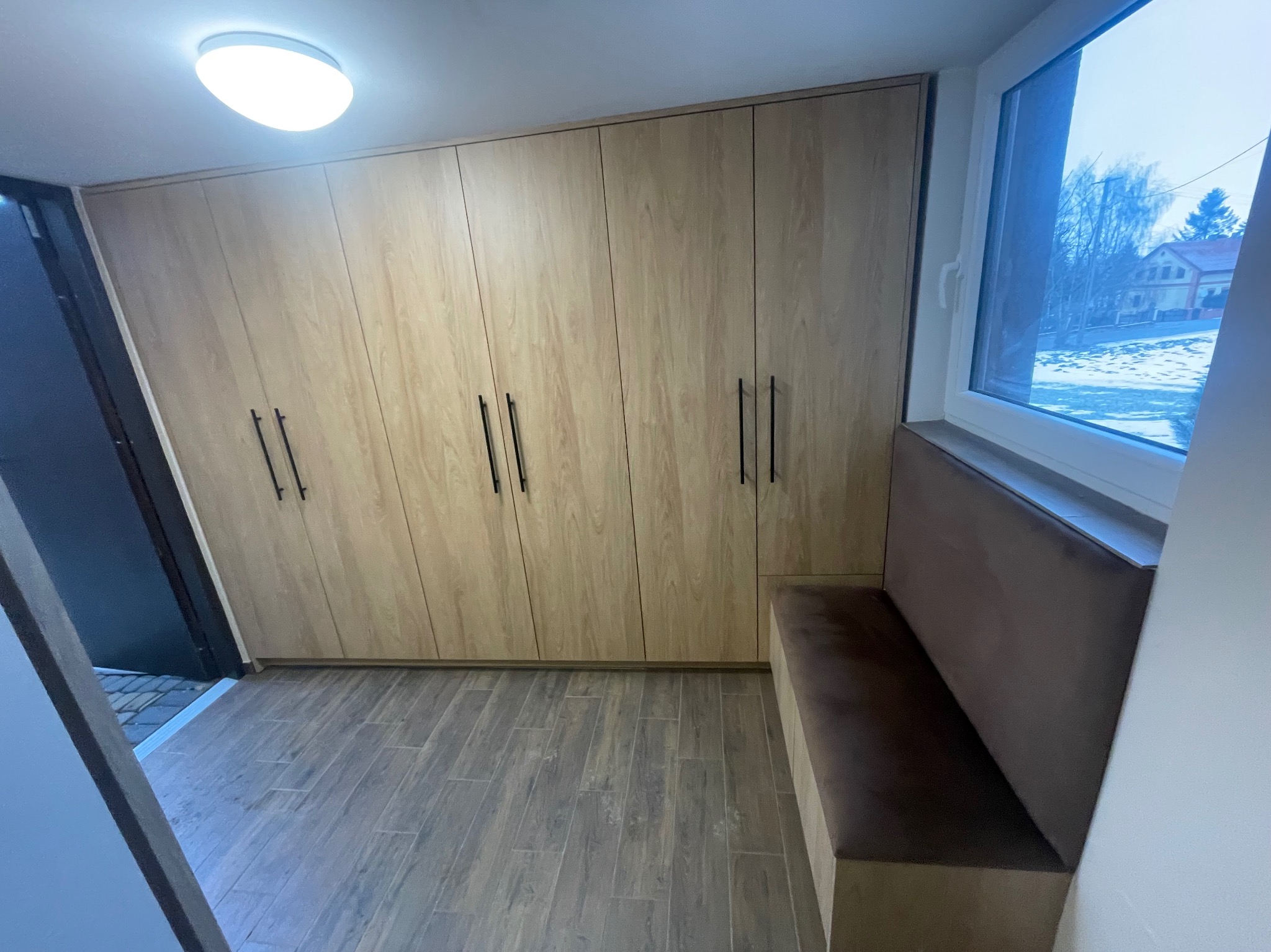 Szafa wnękowa z drewnianymi frontami i czarnymi uchwytami, zintegrowana z siedziskiem pod oknem. Widok z perspektywy wejścia, podłoga z płytek imitujących drewno.