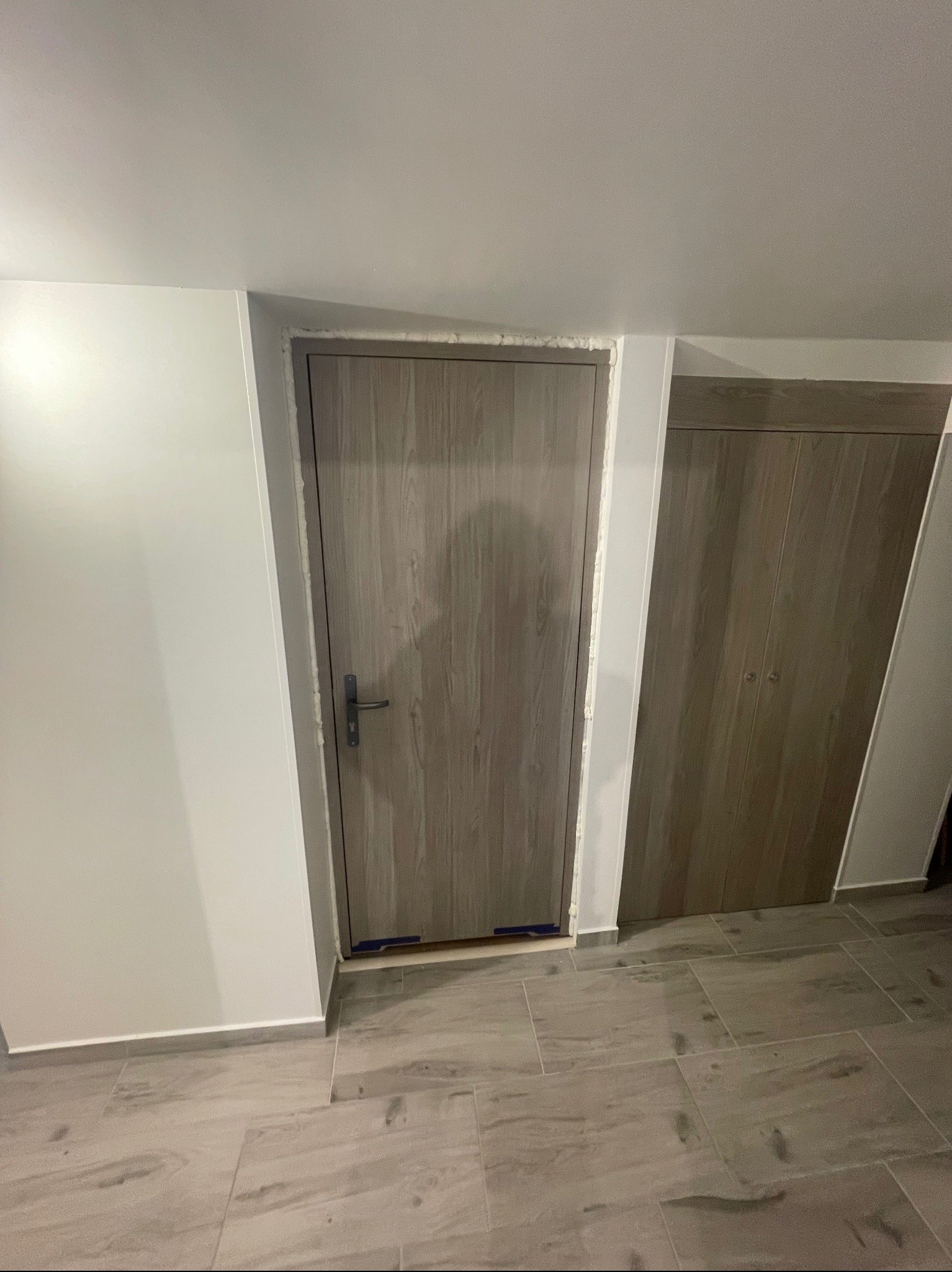 Zamontowane drzwi wewnętrzne z ościeżnicą, widoczna pianka montażowa. Obok szafa wnękowa z drewnopodobnymi frontami. Podłoga wyłożona płytkami imitującymi drewno.