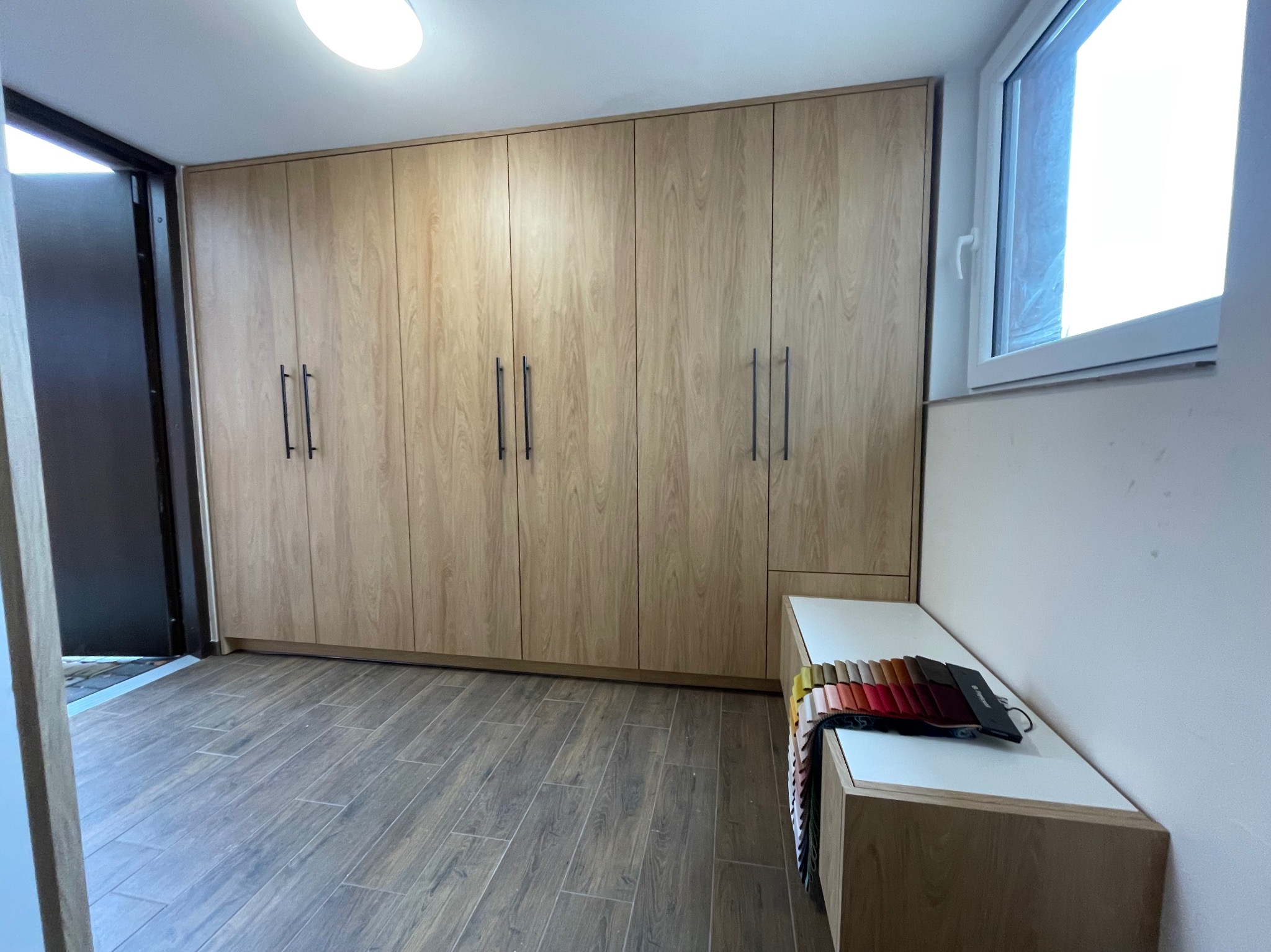 Jasna szafa na wymiar z drewnianymi frontami i czarnymi uchwytami, dopasowana do wnętrza. Na blacie próbki materiałów. Widok z perspektywy podłogi.