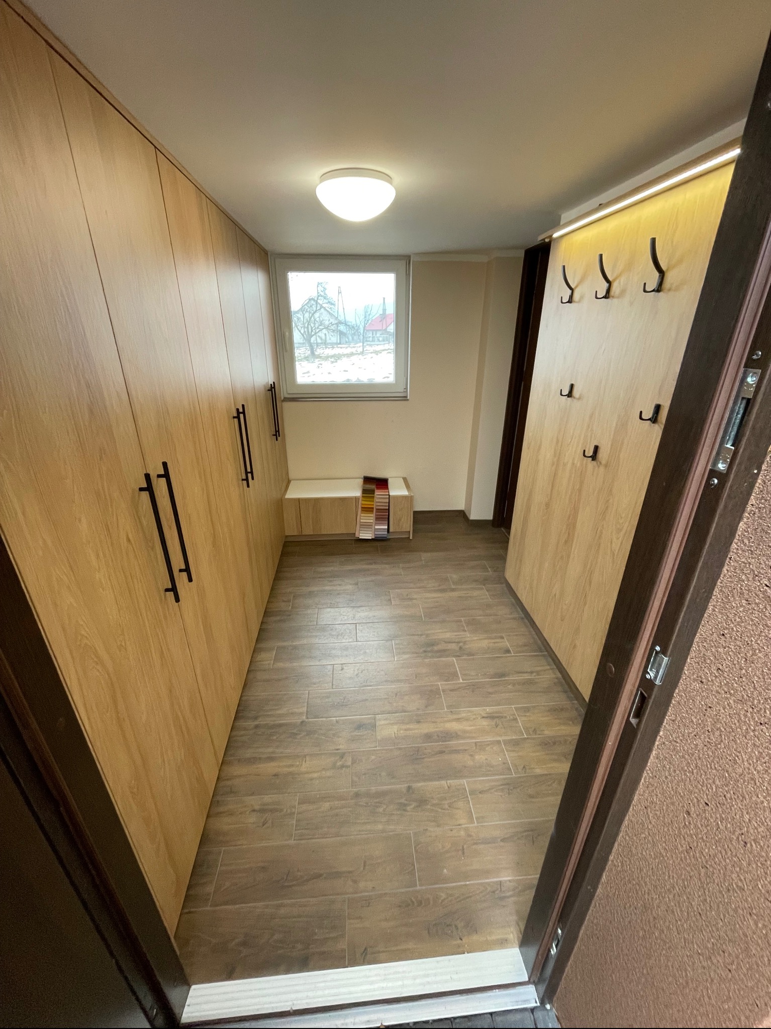 Wnętrze przedpokoju z zabudową szaf w odcieniach drewna, czarne uchwyty, siedzisko pod oknem, wieszaki na ścianie, podłoga imitująca drewno, widok zza otwartych drzwi.