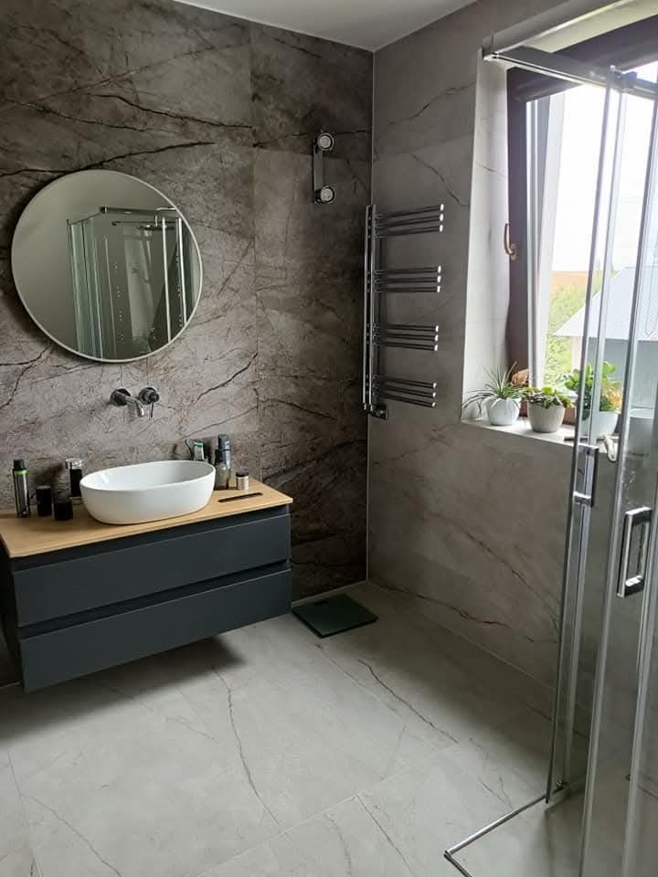 Jasna łazienka z marmurowymi płytkami, okrągłym lustrem i szklaną kabiną prysznicową. Umywalka nablatowa na szafce z drewnianym blatem. Grzejnik drabinkowy.