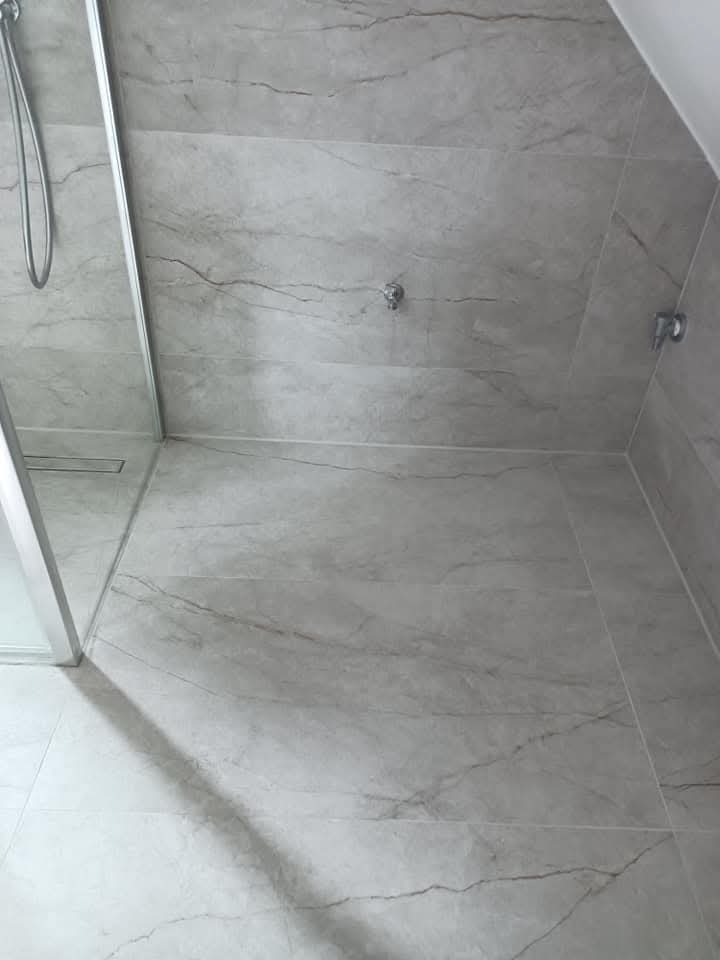 Wykończona kabina prysznicowa z szarymi płytkami imitującymi marmur na ścianach i podłodze. Widoczny odpływ liniowy i elementy armatury.