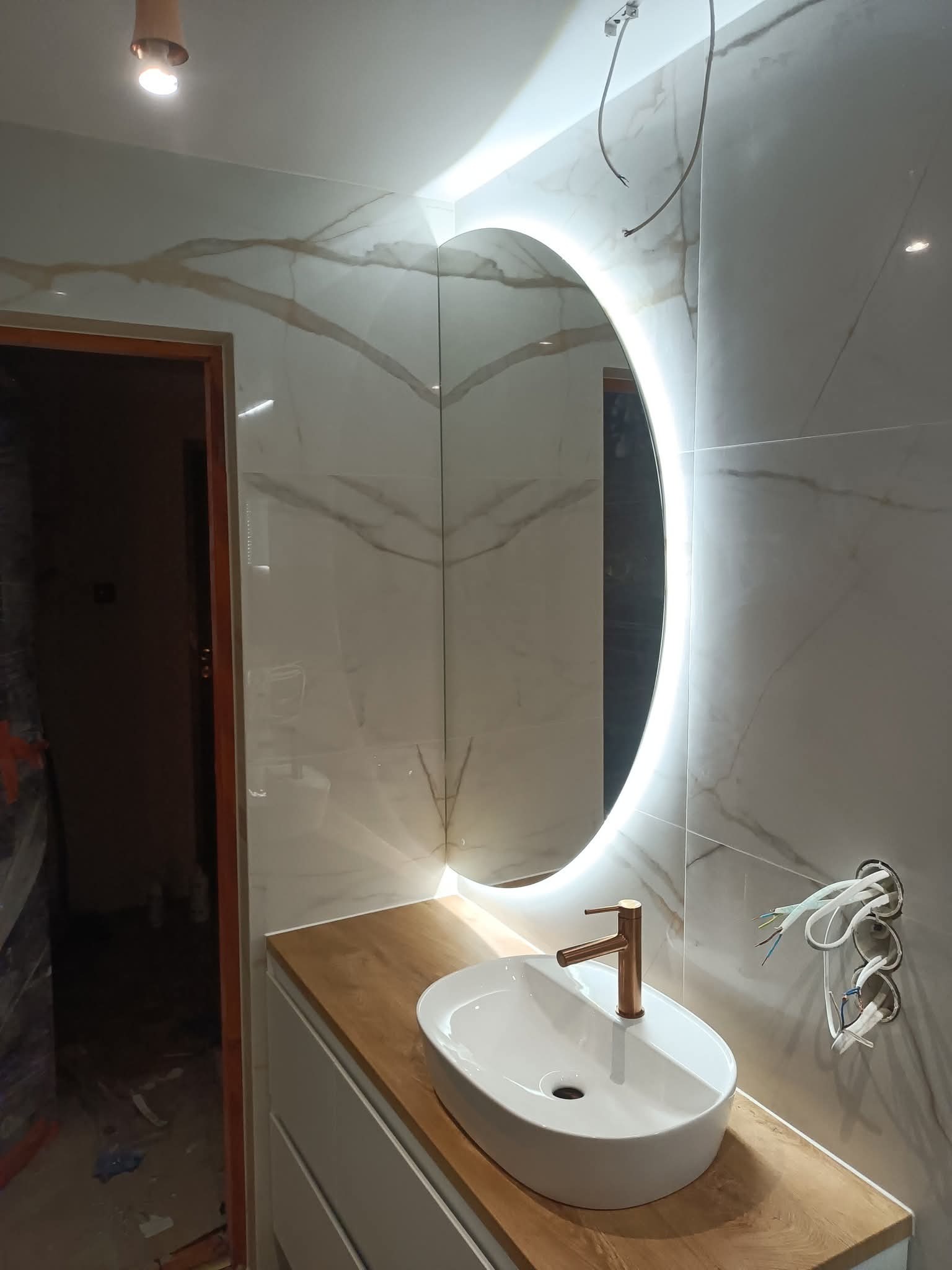 Nowoczesna łazienka w trakcie wykańczania: okrągłe lustro z podświetleniem LED, biała umywalka nablatowa z miedzianą baterią na blacie z drewnopodobnego materiału.