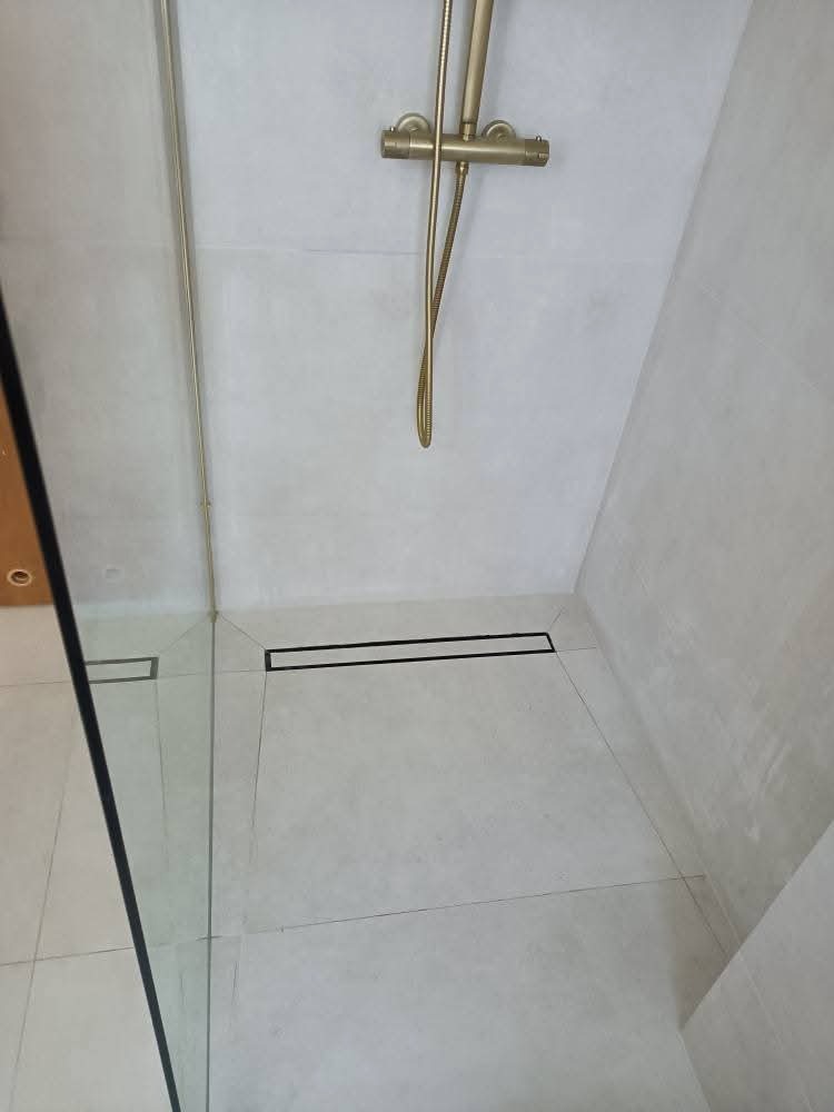 Wykończona kabina prysznicowa z odpływem liniowym, złota armatura i jasne płytki. Minimalistyczny design.