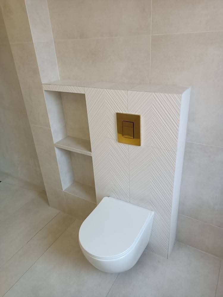 Nowoczesna toaleta podwieszana z białym sedesem i zabudową z półkami, wykończona płytkami w jodełkę. Złoty przycisk spłukujący dodaje elegancji.