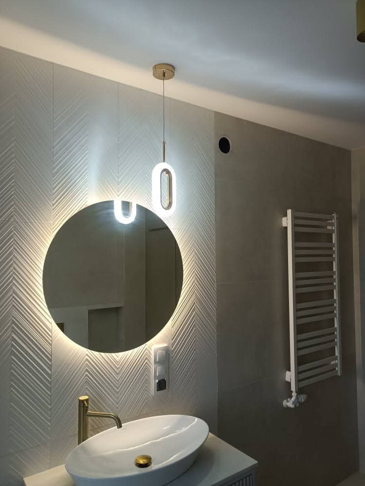 Nowoczesna łazienka z okrągłym lustrem LED, białą umywalką nablatową i grzejnikiem drabinkowym. Ściana wyłożona płytkami w jodełkę. Minimalistyczny design.