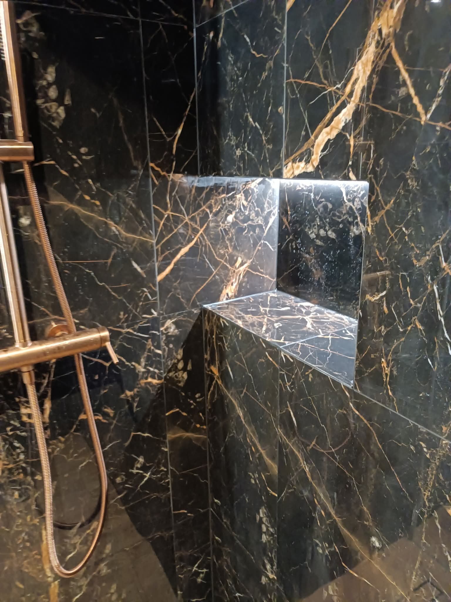 Elegancka, czarna łazienka z złotymi żyłkami na płytkach. Wnęka w ścianie z podświetleniem LED. Elementy armatury w kolorze różowego złota.