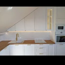 Biała kuchnia z drewnianym blatem, złotymi uchwytami i oświetleniem LED pod szafkami wiszącymi, dopasowana do skosu dachu, z białymi płytkami na ścianie.