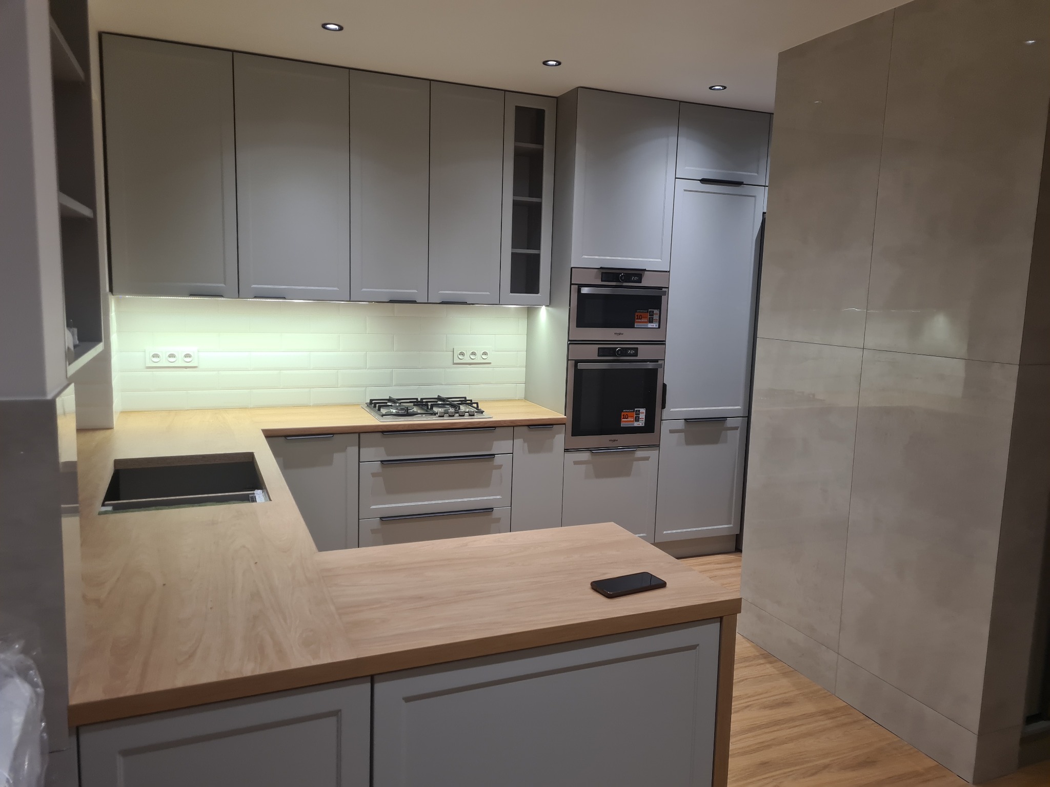 Jasnoszara kuchnia z drewnianym blatem, widoczny zlew i płyta gazowa, na blacie leży smartfon