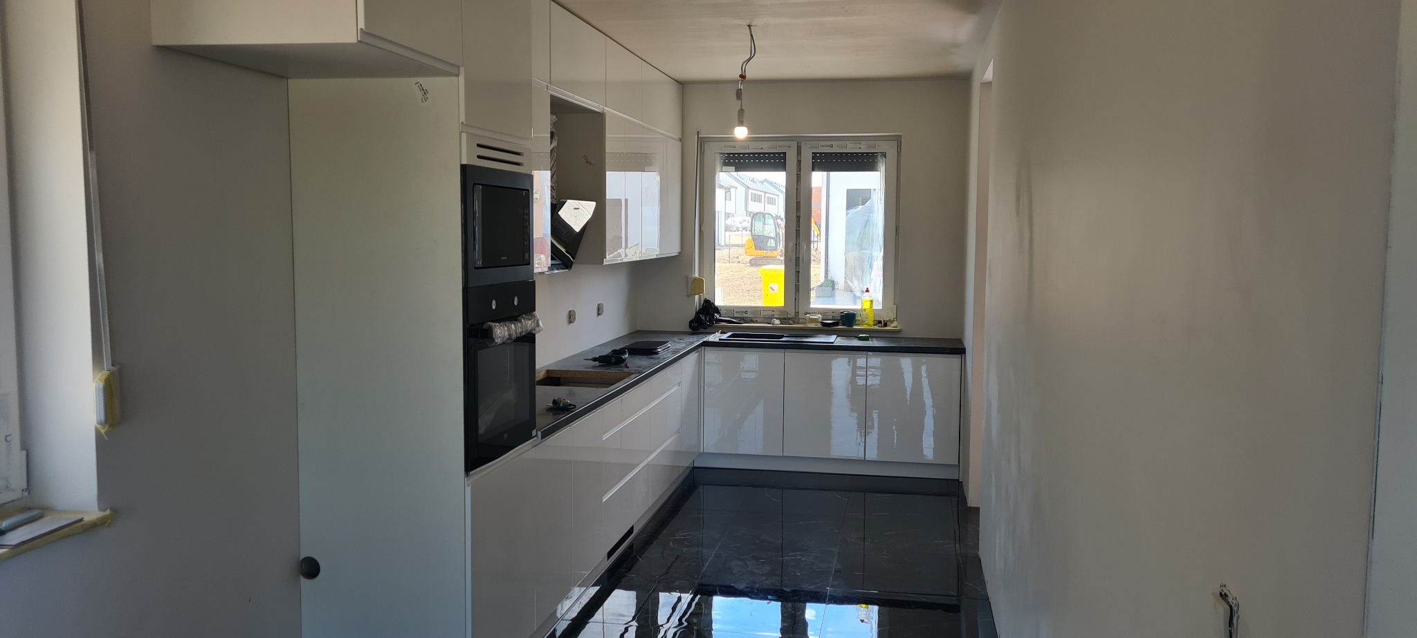 Wnętrze kuchni w trakcie montażu z białymi, lakierowanymi frontami, czarnym blatem i błyszczącą podłogą; widoczny piekarnik i mikrofalówka w zabudowie, a przez okno koparka na zewnątrz budynku.