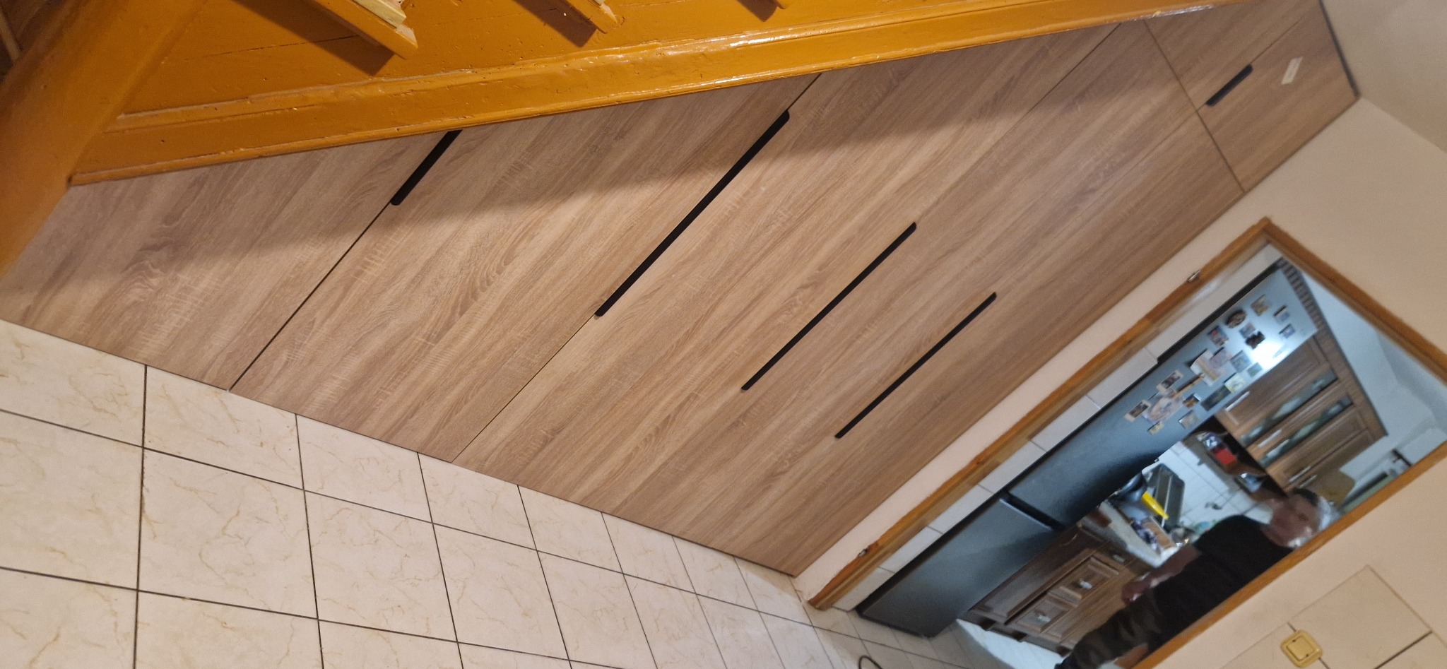 Zabudowa kuchenna z frontami z płyty drewnopodobnej z ukośnymi uchwytami, widok od dołu, fragment podłogi z płytek.