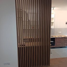 MaTo Meble - Nowoczesna, drewniana ścianka działowa z pionowych listew oddzielająca kuchnię. Widoczny fragment zabudowy kuchennej w tle. Minimalistyczny design, ciepłe oświetlenie.