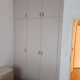 MaTo Meble - Jasnoszara szafa wnękowa z drewnianymi uchwytami, zabudowana od podłogi do sufitu, widoczna z boku w pokoju z drewnianą podłogą i fragmentem łóżka.