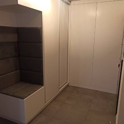 MaTo Meble - Biała szafa wnękowa z tapicerowanym siedziskiem i oświetleniem LED u góry. Minimalistyczny design, szara podłoga, drzwi przesuwne.