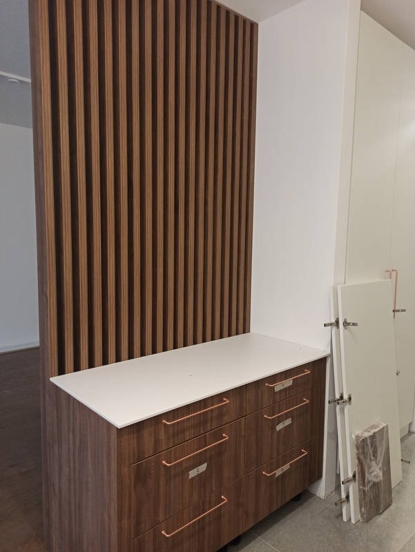 Nowoczesna komoda z drewnianymi frontami i białym blatem, uzupełniona o pionowe panele dekoracyjne. Widoczne drzwi szafek do montażu, sugerujące etap wykończenia.