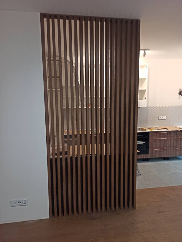 Nowoczesna, drewniana ścianka działowa z pionowych listew oddzielająca kuchnię. Widoczny fragment zabudowy kuchennej w tle. Minimalistyczny design, ciepłe oświetlenie.